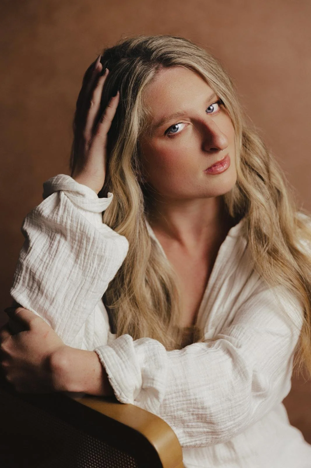 A young woman with long blonde hair and blue eyes is resting her head on her hand, wearing a white textured blouse, against a warm brown background.