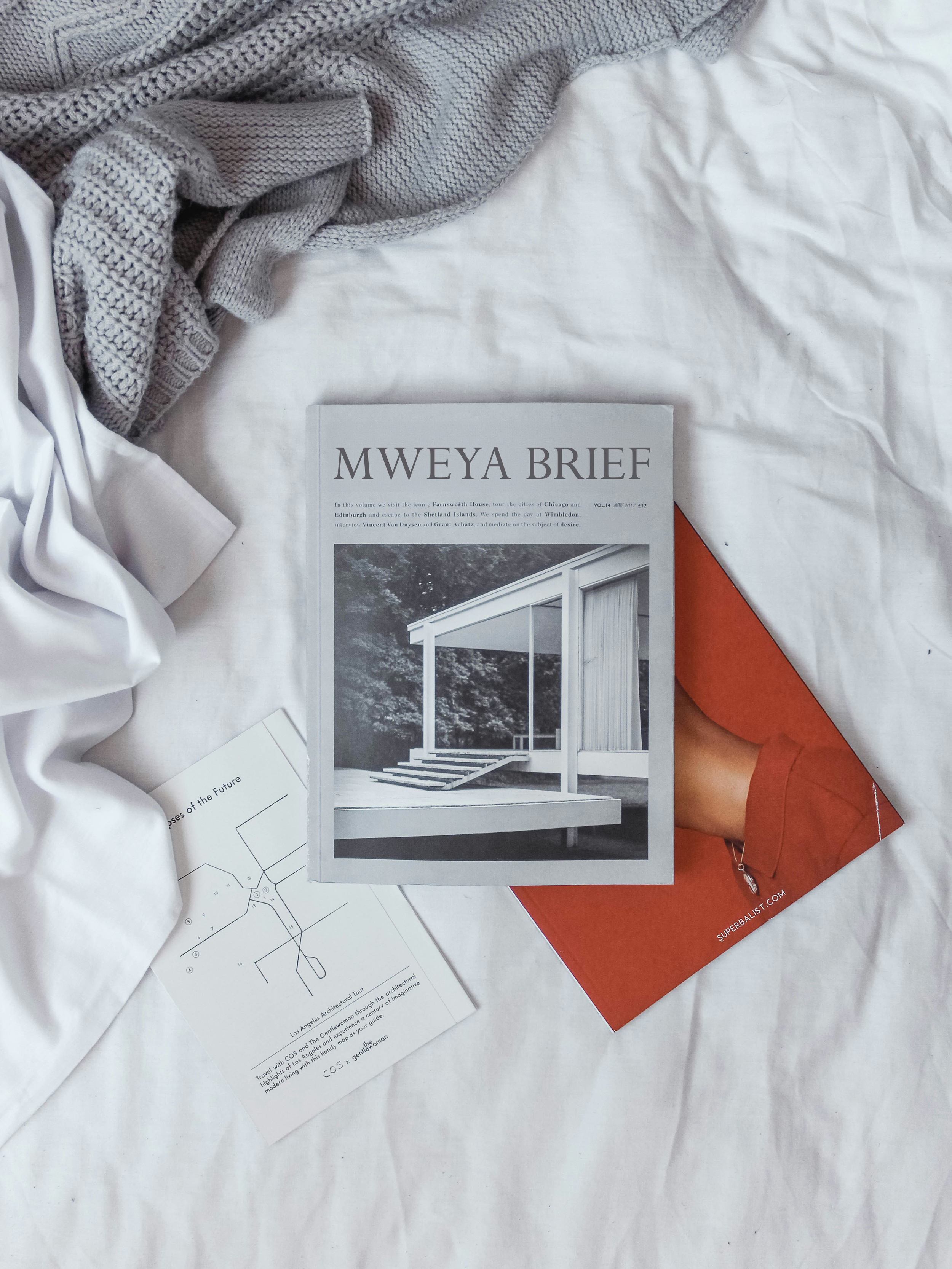Magazine titled "MWEYA BRIEF" on a bed with a white sheet, surrounded by gray knitted and woven blankets, with a red magazine partially visible underneath.