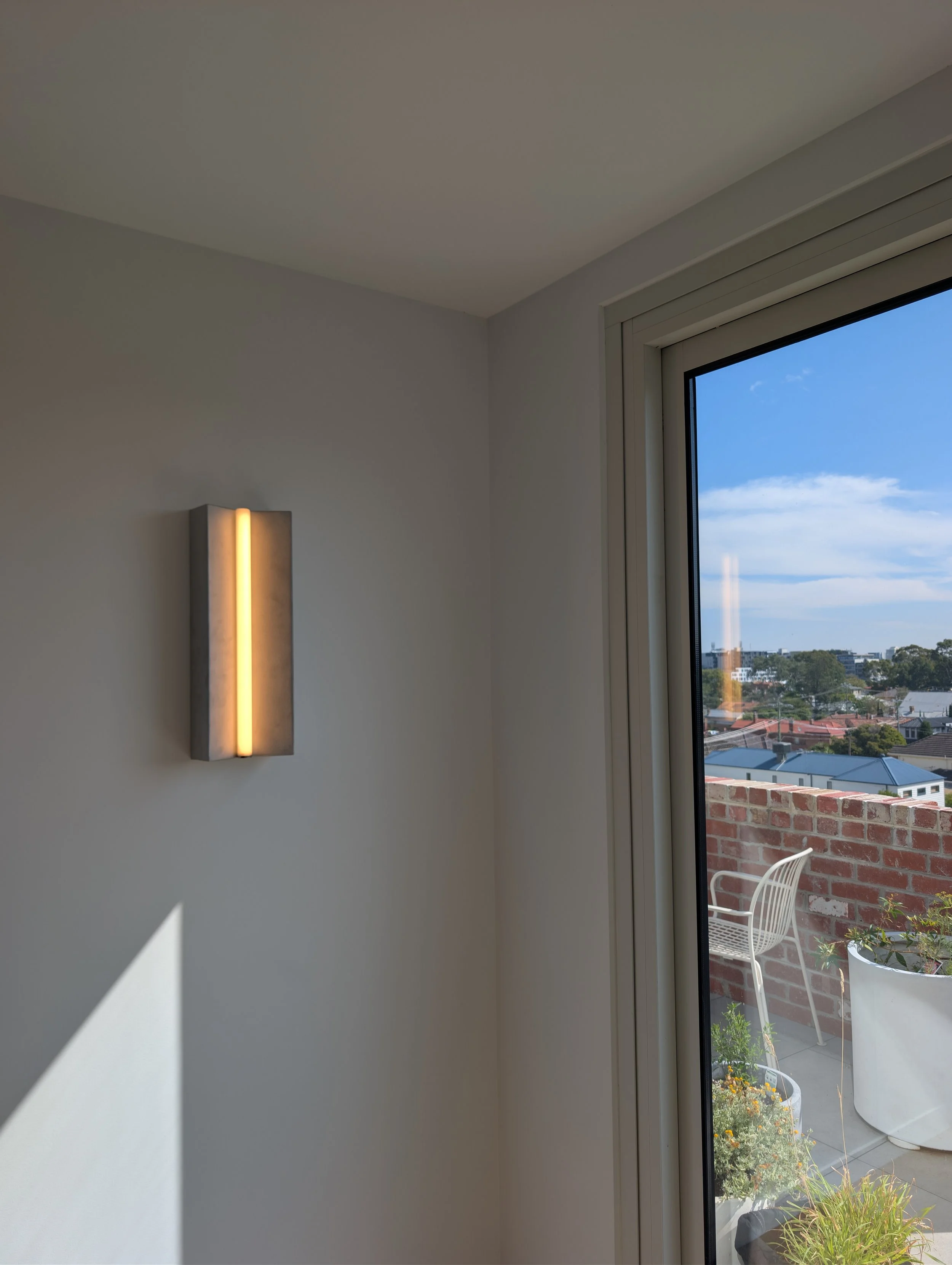 A corner of a room with a modern wall-mounted light fixture and a large window showing a cityscape with trees, buildings, and a fenced patio with outdoor furniture and potted plants.
