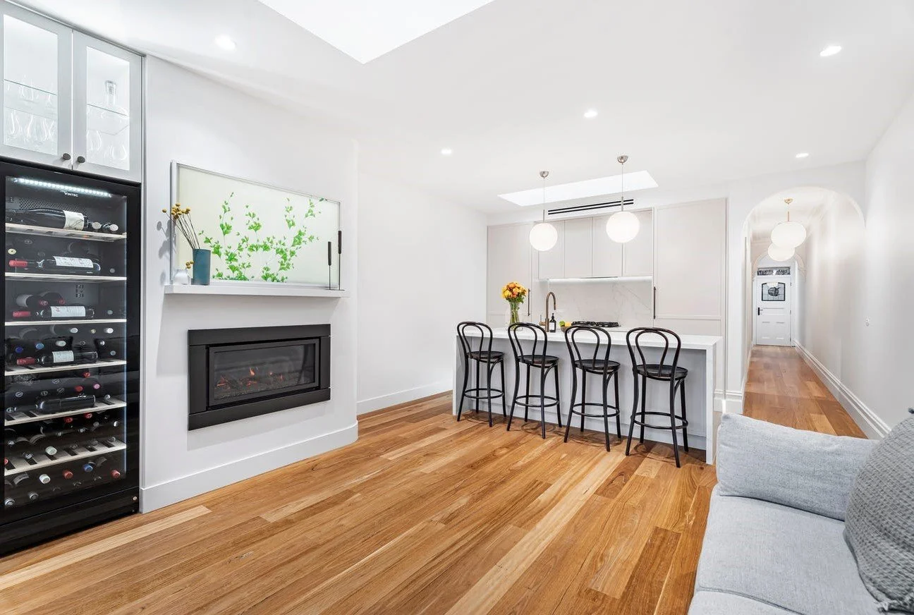 Modern open-concept living space with a kitchen island, four barstools, a wine fridge, a fireplace with a glass wall, and wooden flooring, illuminated by ceiling lights and pendant lights.