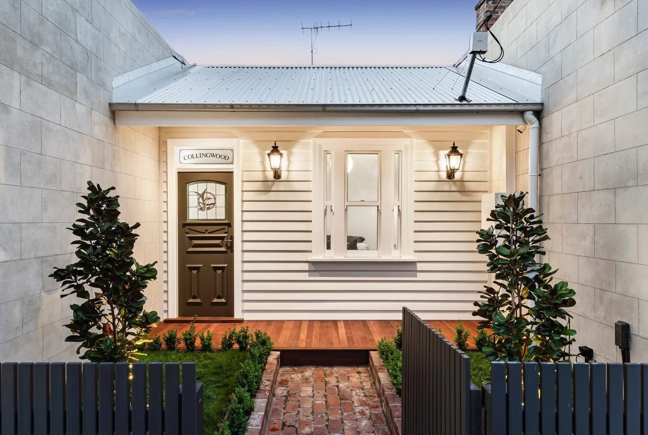 Front porch of a house with a black front door, white exterior walls, a small window, and two wall-mounted lanterns. The porch has a wooden platform and brick pathway, with two tall shrubs and smaller plants on either side, surrounded by a black fenc