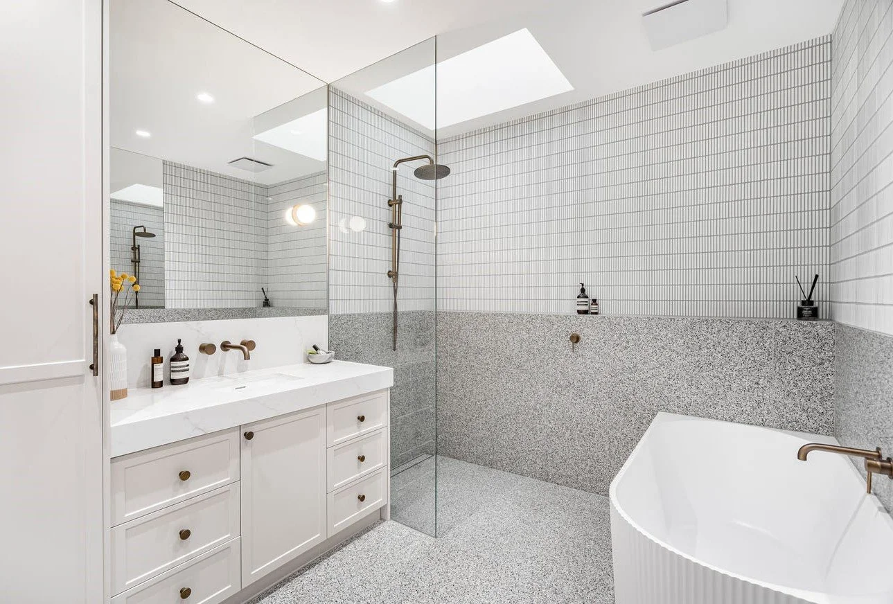 Modern bathroom with white vanity, large mirror, terrazzo flooring, and walk-in shower with skylight and a separate bathtub.