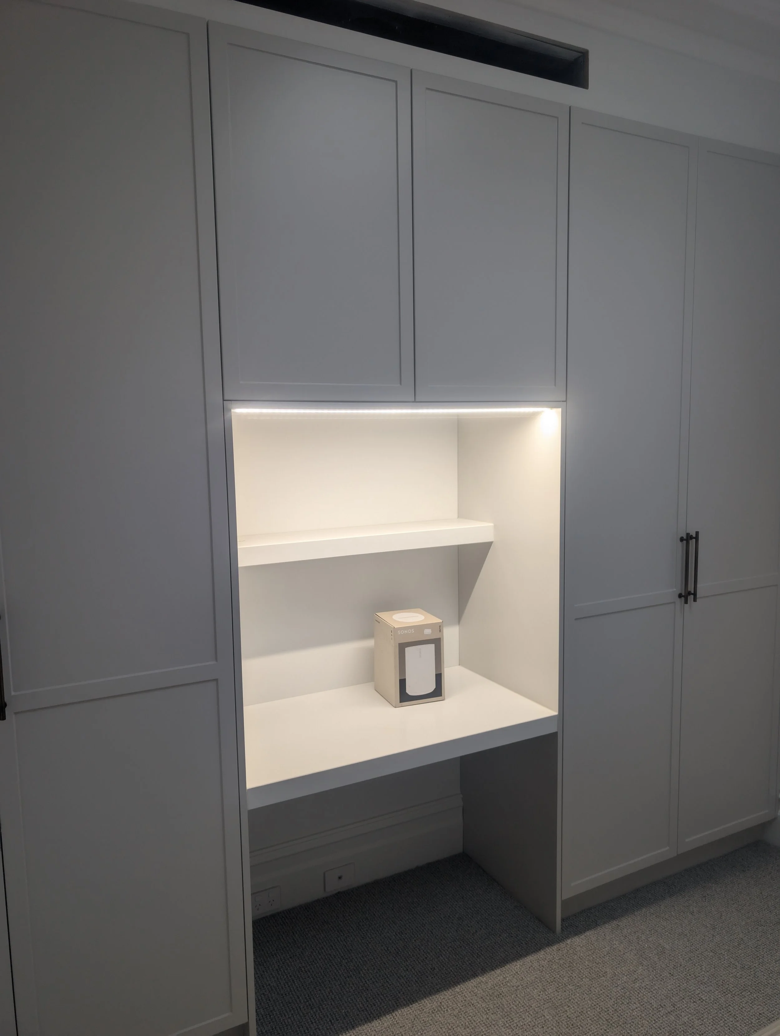 Built-in white cabinets with closed doors surrounding an open shelf with built-in lighting. A boxed product with 