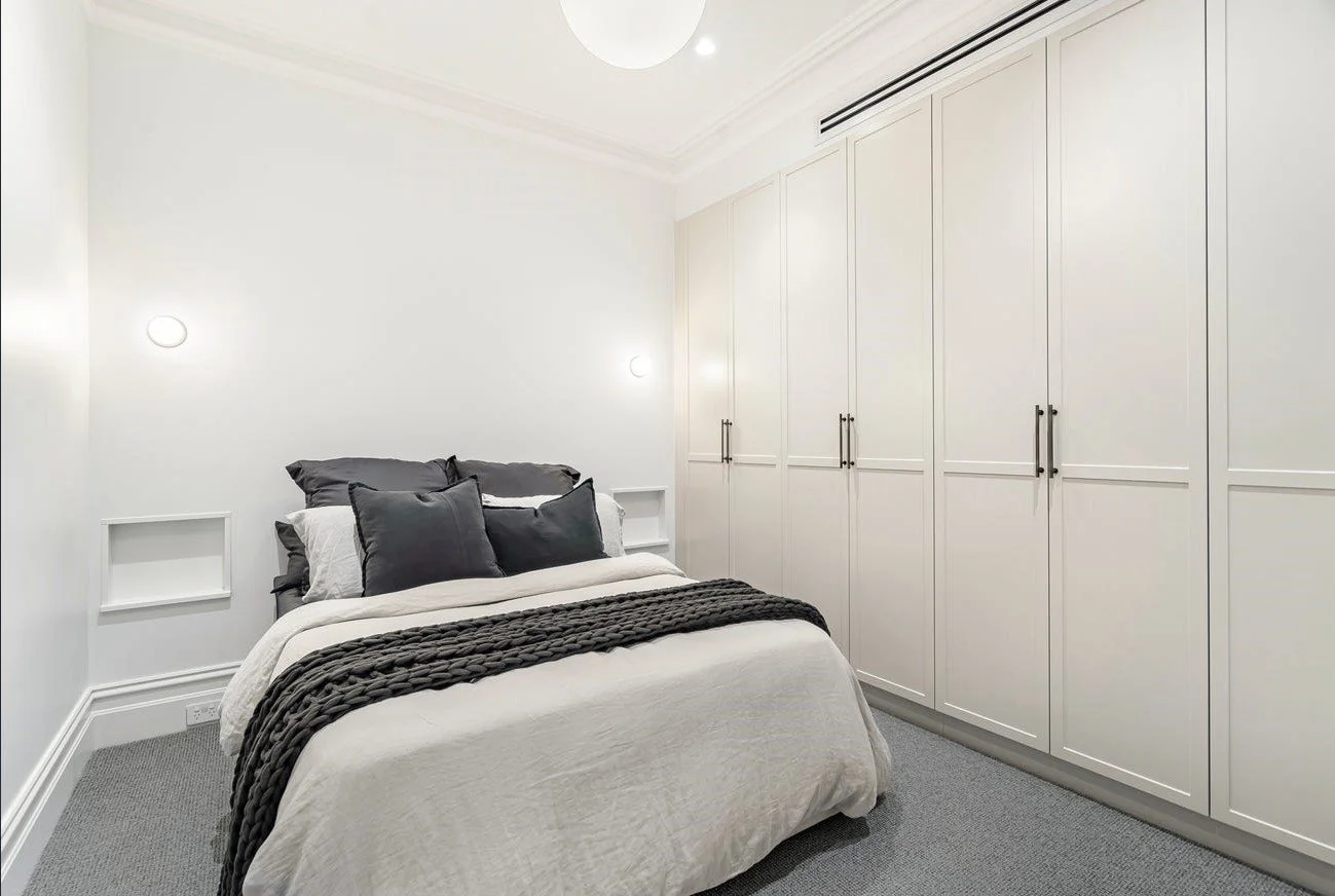 A neatly made bed with dark gray pillows and a chunky knit throw blanket in a minimalist white bedroom with built-in white closet doors and soft lighting.