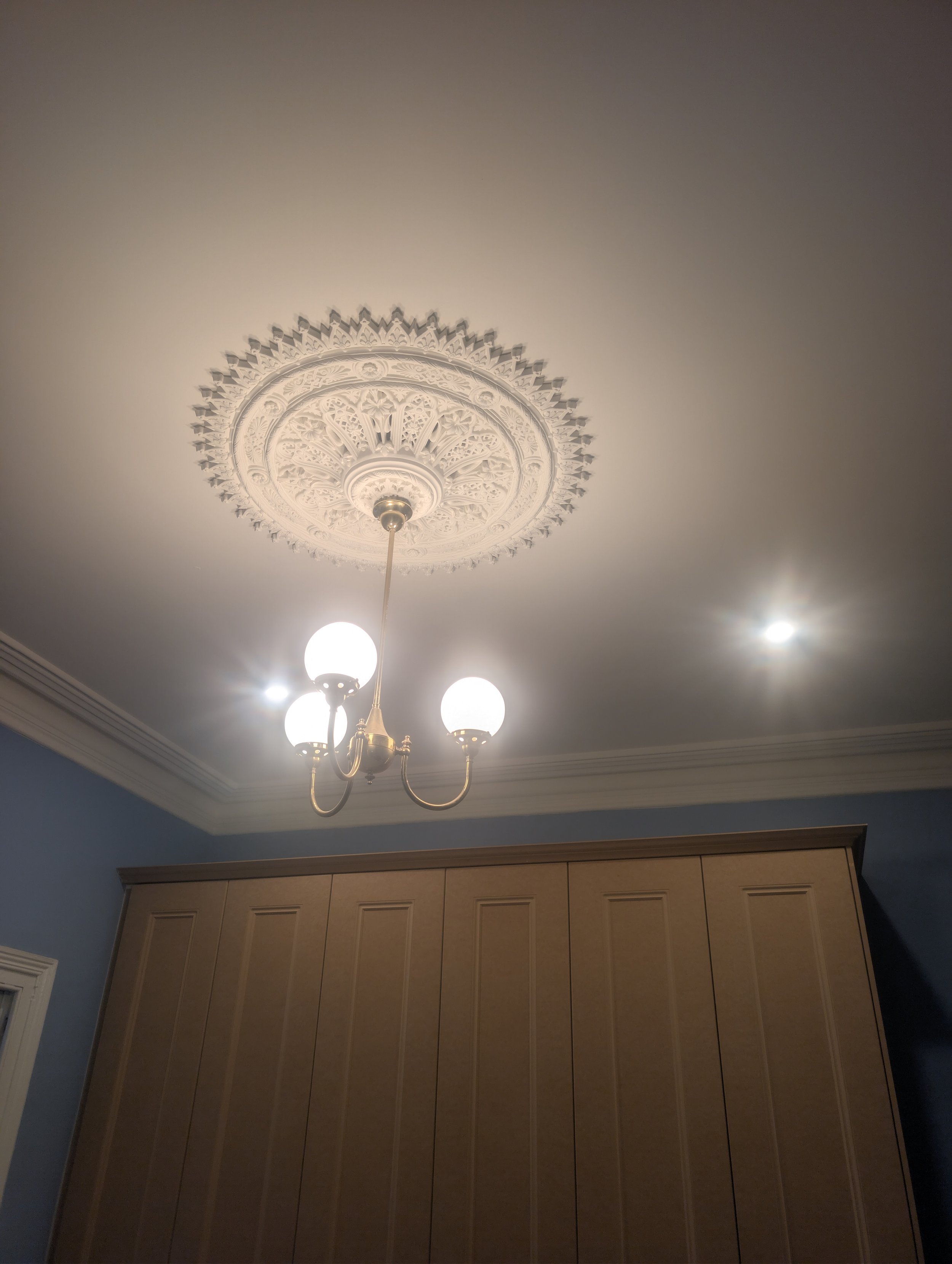 Ceiling with decorative medallion and hanging chandelier with five globe-shaped bulbs in a room with beige walls and wooden cabinets.