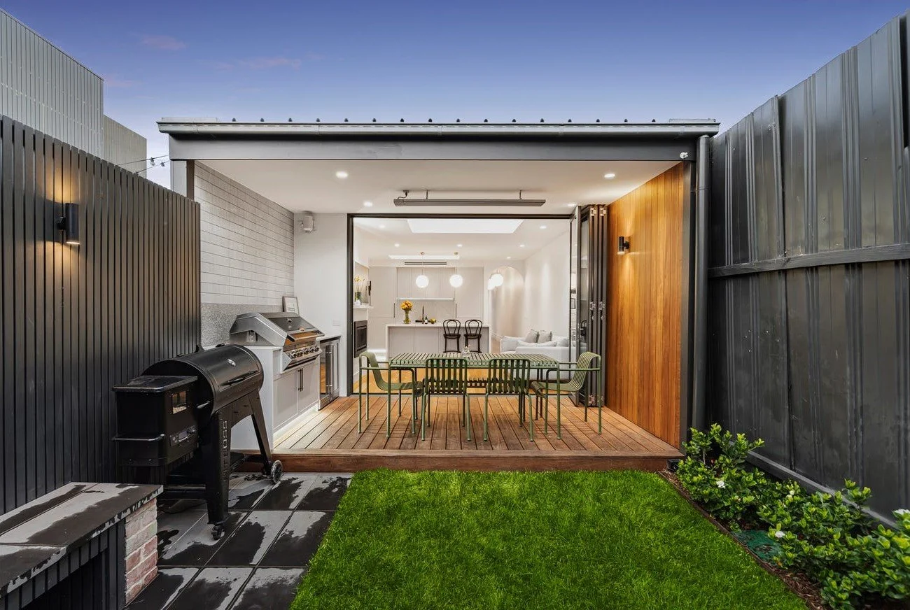 Modern outdoor patio with a green grassy yard, black wooden fence, and a covered dining area with a table and green chairs. The patio has a built-in grill and outdoor light fixtures, overlooking an indoor living space with a kitchen and seating area.