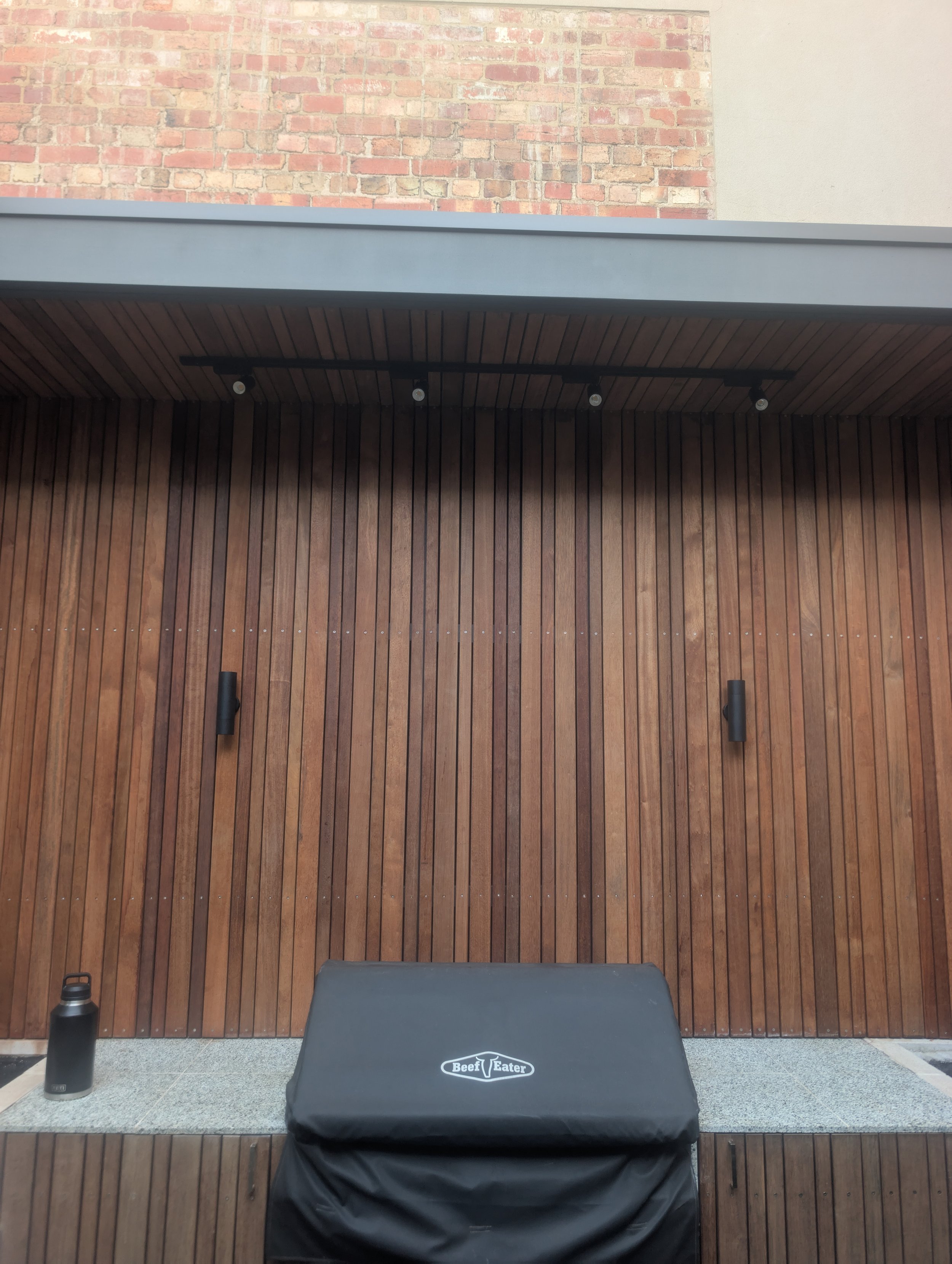 Outdoor patio area with wooden panel wall, a black table covered with a BeefEater cover, and a black water bottle on a gray surface, brick wall in the background.