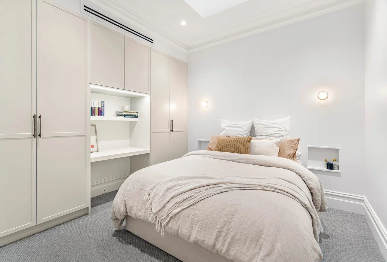 A modern, minimalist bedroom with a white bed, beige bedding, and built-in cabinetry on one wall, illuminated by wall sconces and ceiling light, with small decorative items on built-in shelves.