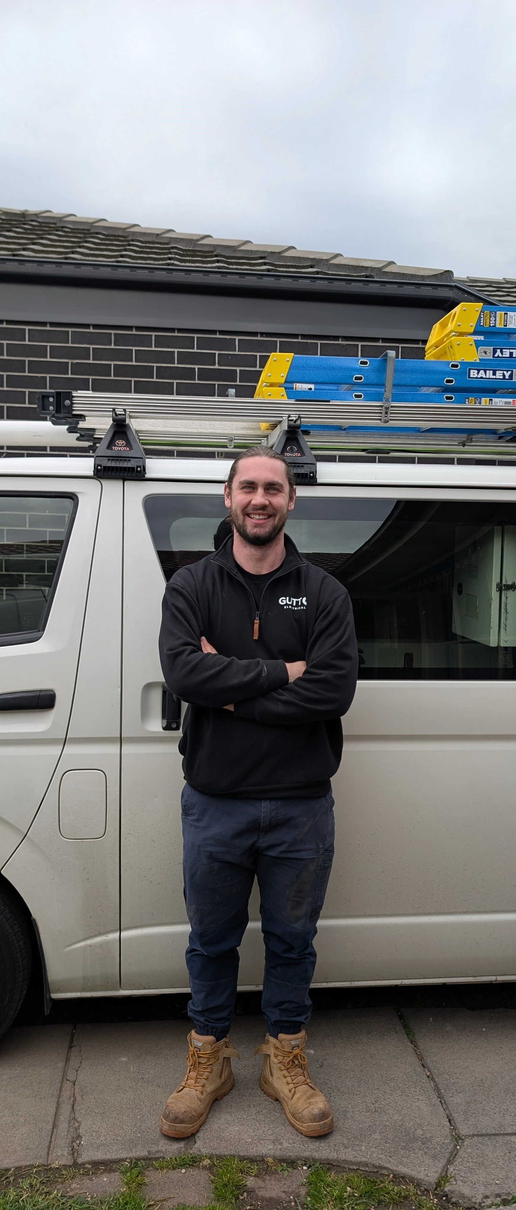 A man standing in front of a white work van with a ladder rack on top, smiling, with a house and cloudy sky in the background.