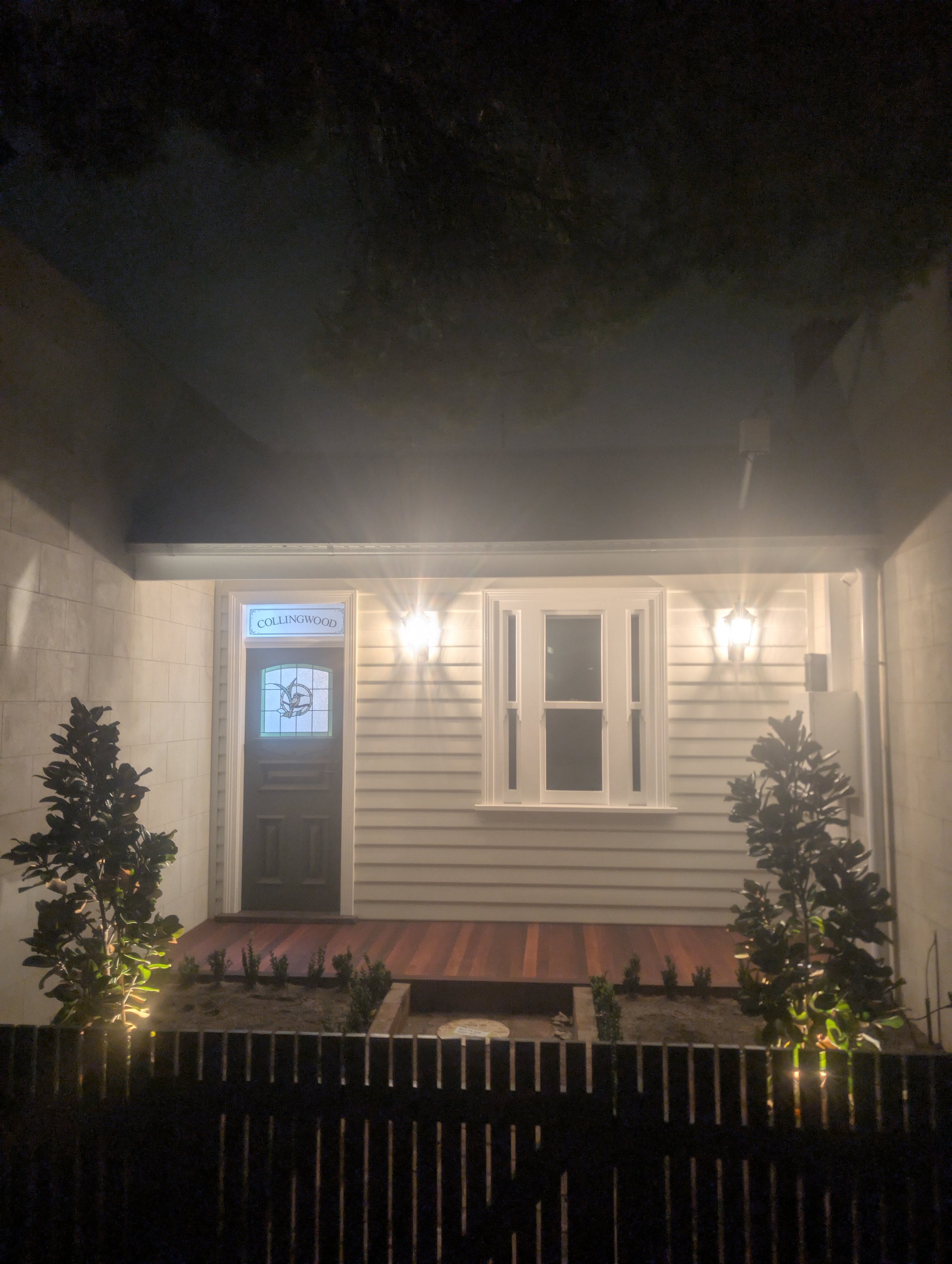Night view of a house entrance illuminated by outdoor lights, with a sign on the window reading 'COLLINGWOOD,' a small front yard with plants, and a black metal fence in the foreground.