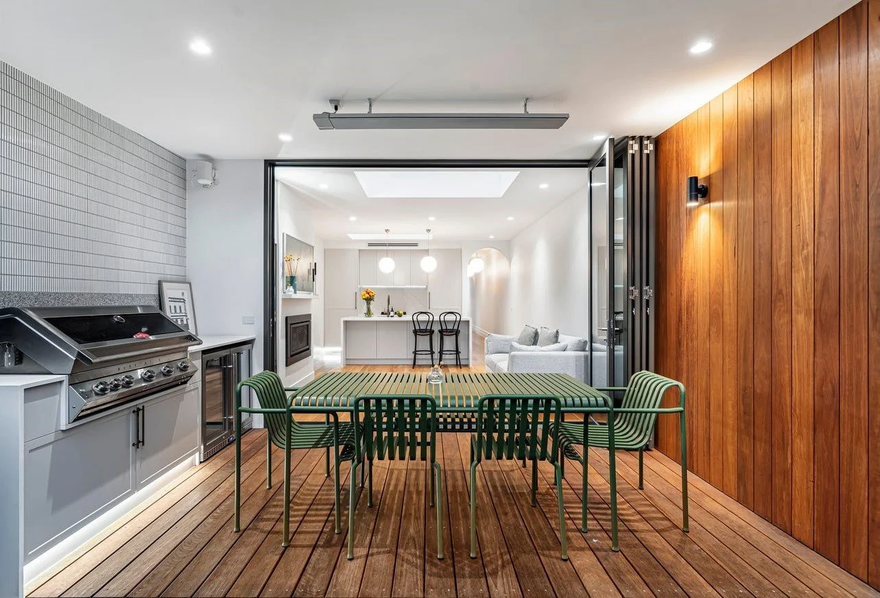 Interior view of a dining area with a wooden table and green chairs, leading to a bright living space with a white sofa and a kitchen.
