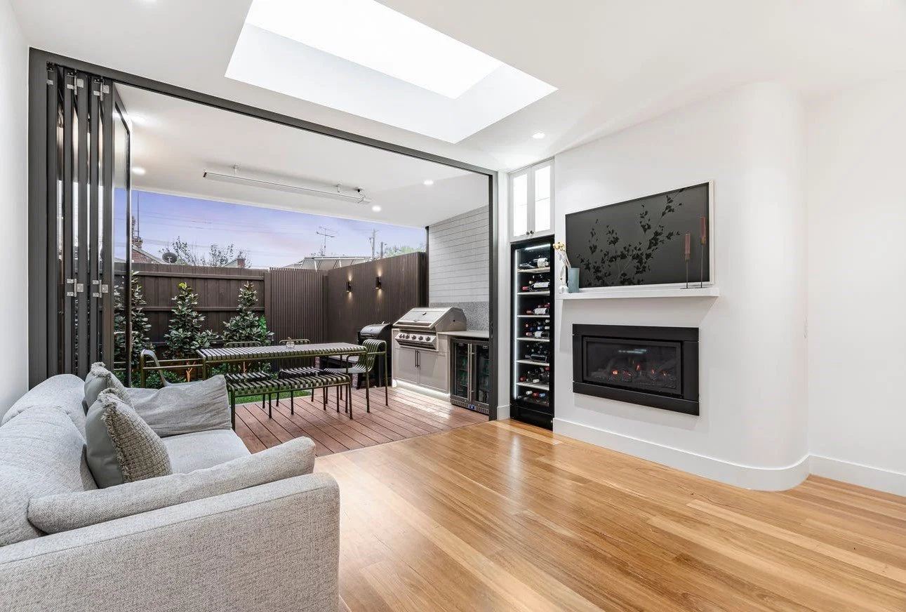 Living room opening to outdoor patio with grill, dining table, and greenery, featuring wood and white interior design, hardwood floors, and wall-mounted TV with fireplace.