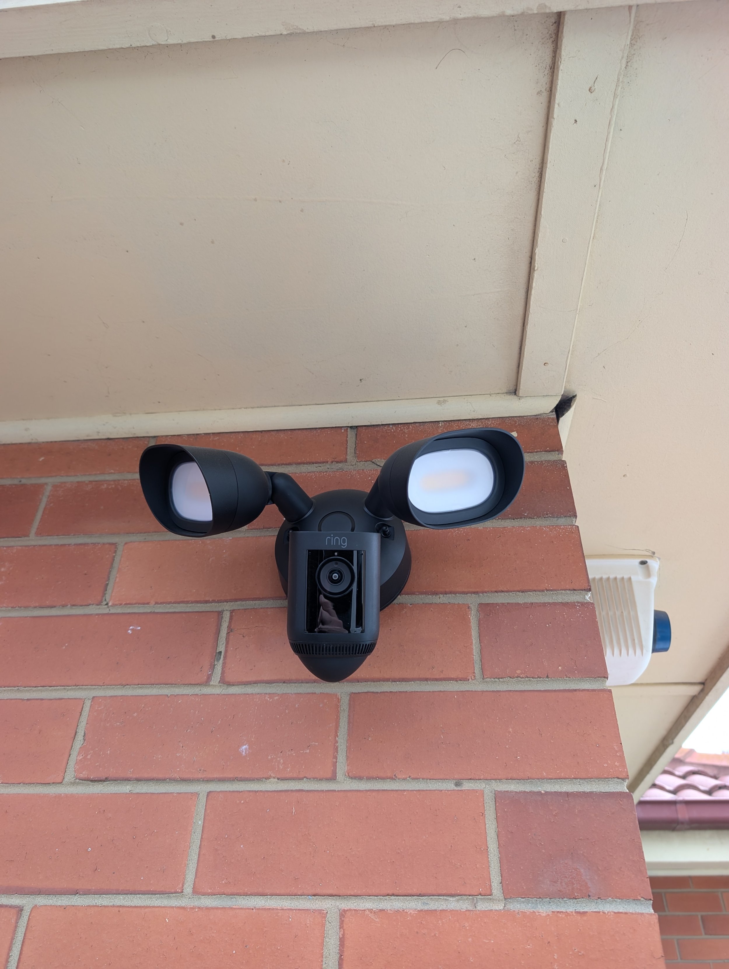 A security camera mounted on a brick wall, with beige panels above and a white vent to the right.