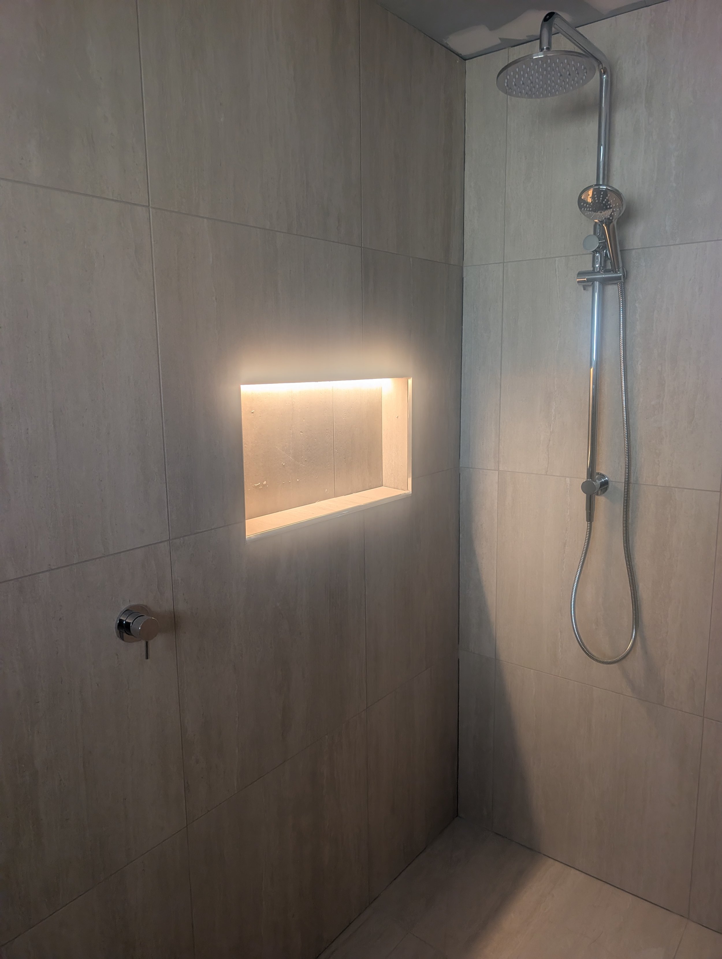 A modern shower with beige tiled walls, a built-in niche with lighting, and a rain showerhead attached to an adjustable bar with a handheld shower. The floor tiles are also beige.