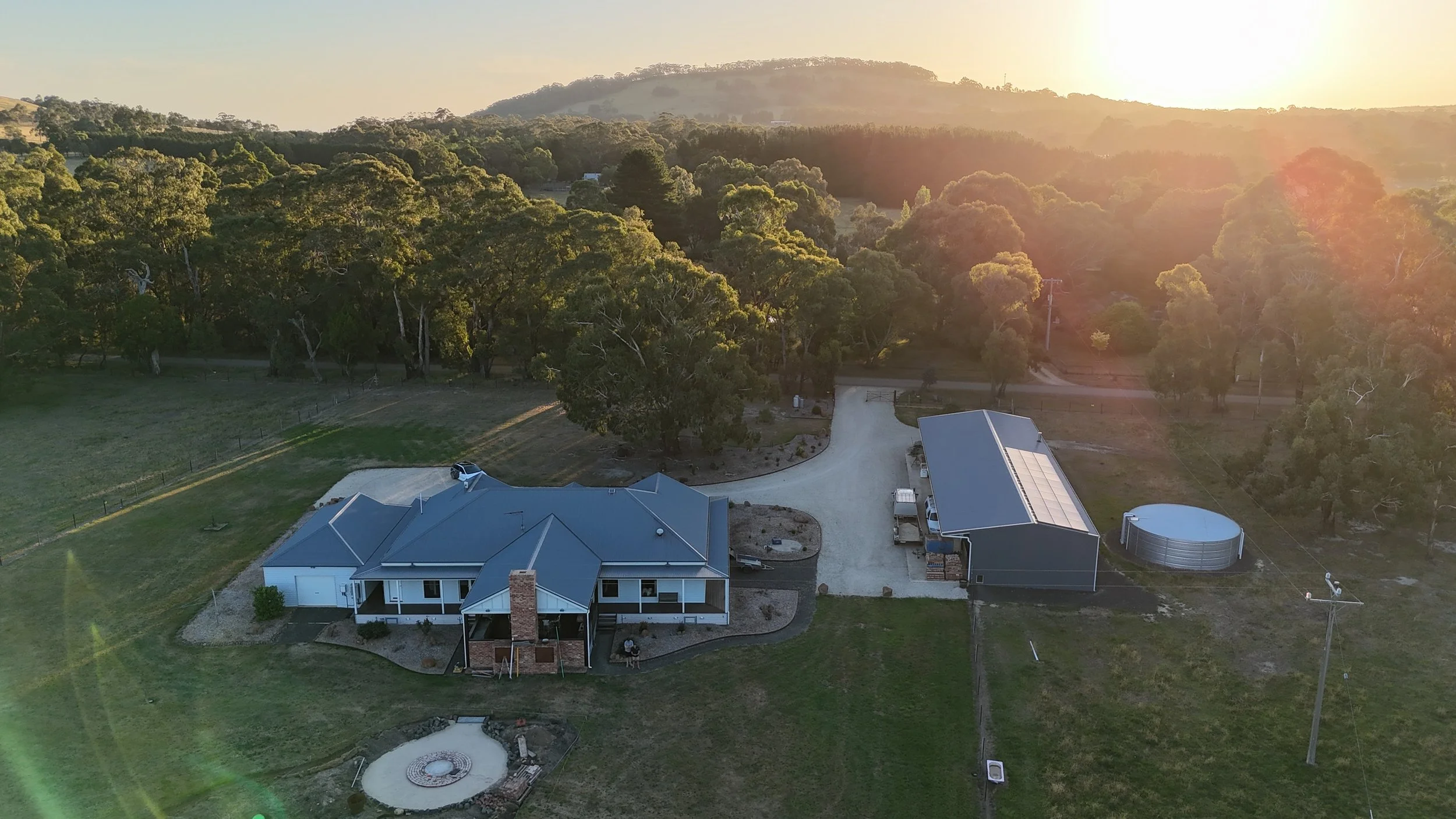 Aerial view of a modern house with a blue roof, surrounded by green lawns, trees, and a driveway, with a mountainous landscape in the background during sunset.