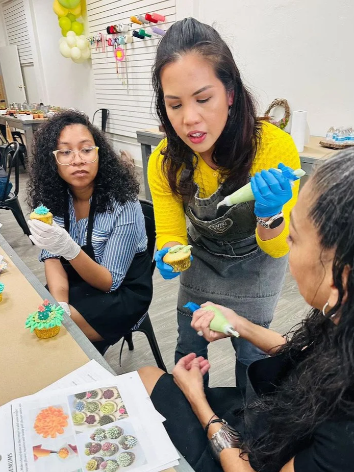 Three women decorating cupcakes in a classroom setting with Joy from MMC Bakes. This is a crafting business, so there are various colorful decorations on the wall.