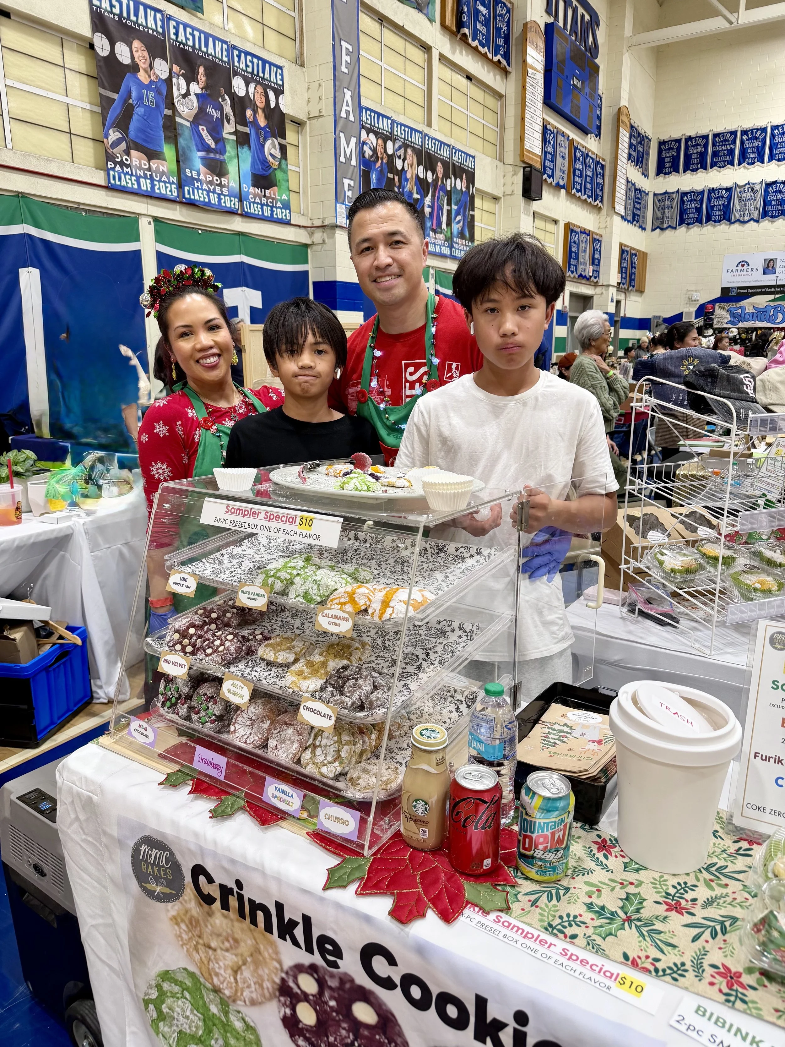 Four people standing behind a table with a display of cookies and snacks at the PASACAT Parol Festival at the Eastlake High School gymnasium in Chula Vista.  The background features banners and posters for Eastlake Titans Volleyball.