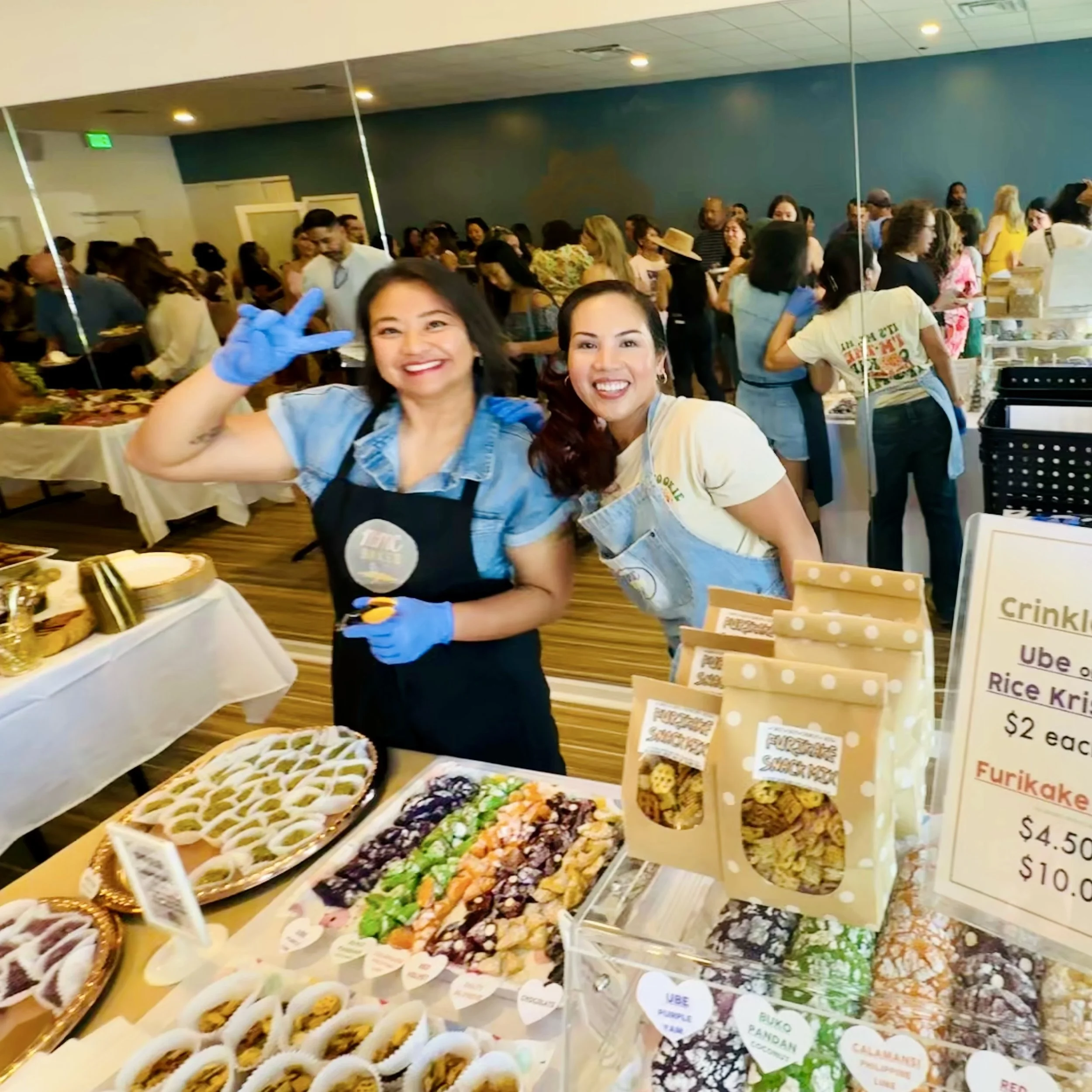 Two women smiling and posing at the Namaste Eastlake grand opening party,  MMC Bakes food stall selling Filipino snacks, and crinkles cookies with a busy crowd and tables in the background.
