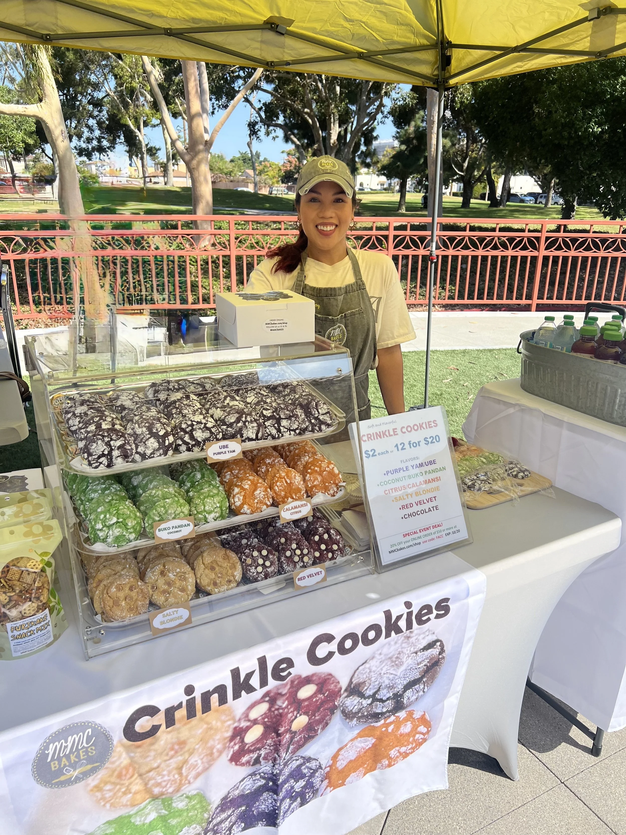 Joy of MMC Bakes smiling, standing behind a table at an outdoor market stall selling crinkle cookies in various flavors. The table has a sign with cookie prices and flavors. This event was the Fil-Am CreatorCon in Chula Vista.