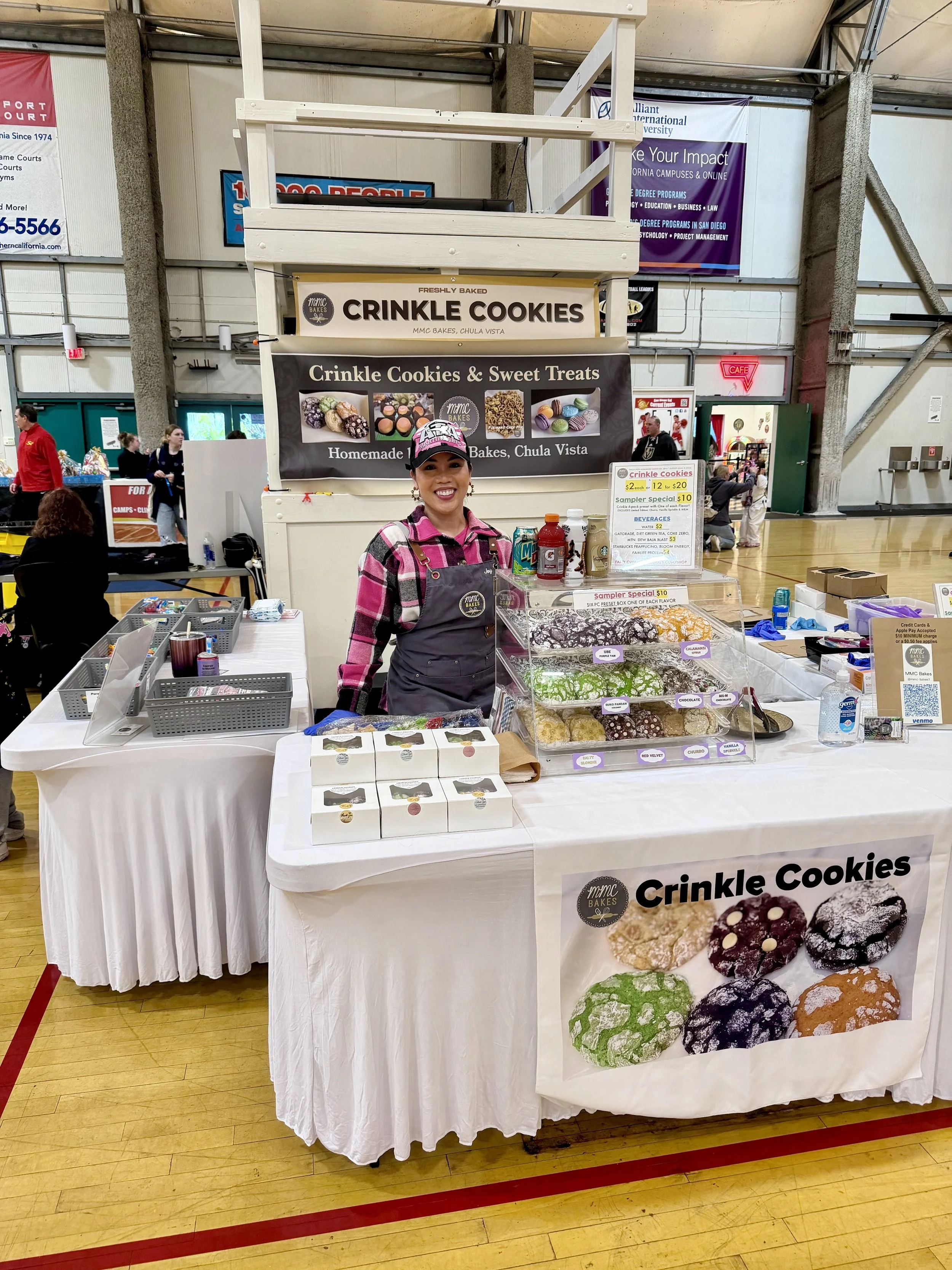 A woman standing behind an MMC Bakes booth selling crinkle cookies at an indoor market, taekwondo tournament. The booth has signs advertising sweet treats, with various cookie boxes and display trays of colorful cookies on the table.