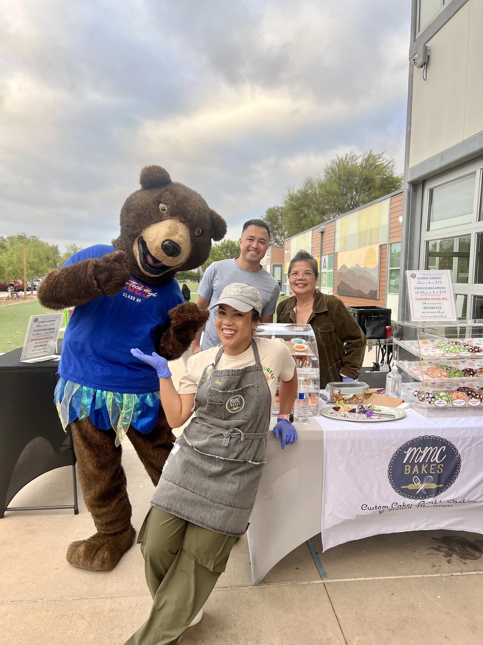 A group of four people and a person in a bear mascot costume are smiling at an outdoor event selling crinkle cookies by MMC Bakes, which was at High Tech High School.