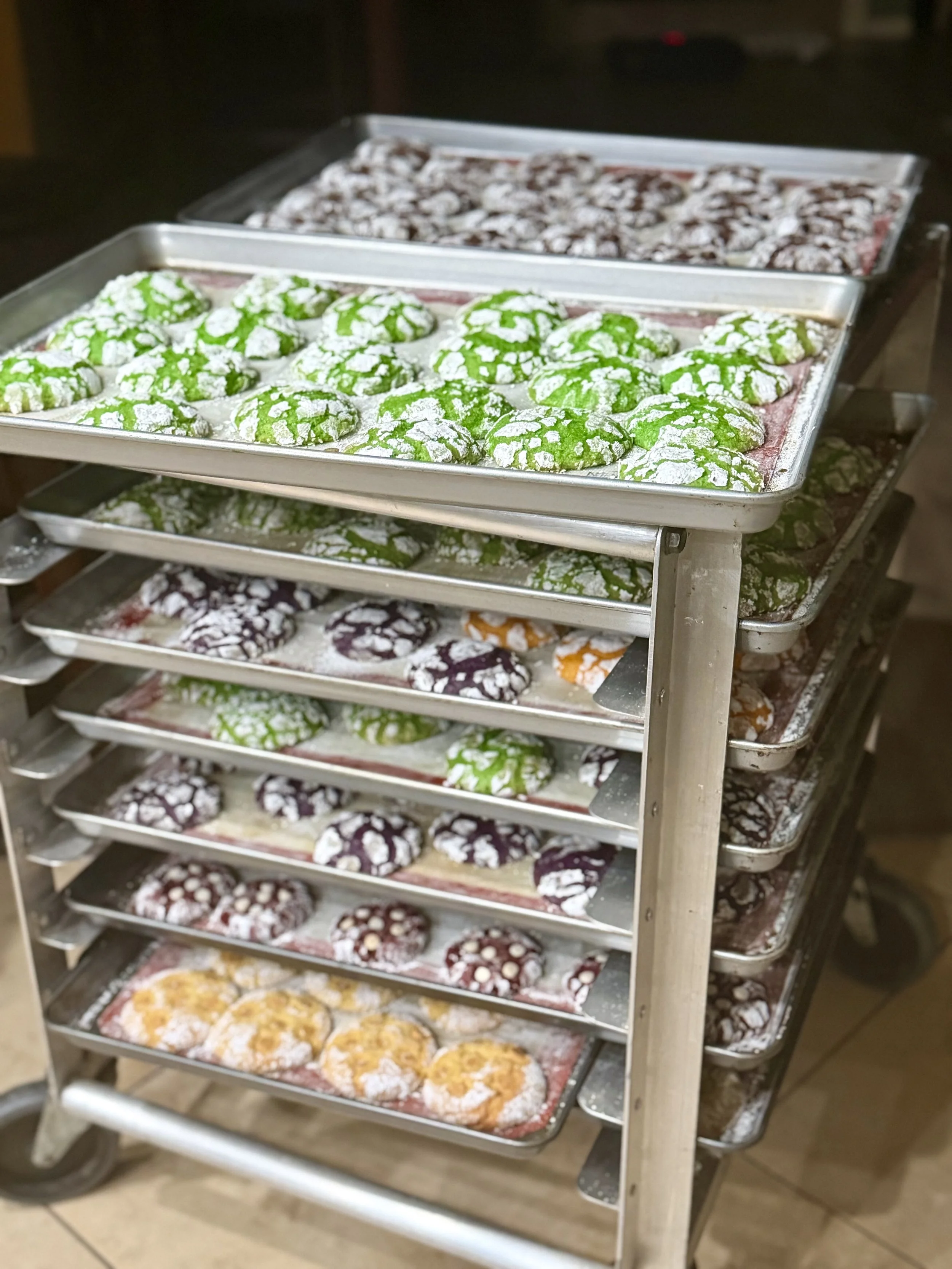 A metal rack with multiple trays of colorful assorted crinkle cookies baked by Joy from MMC Bakes in a home bakery kitchen.