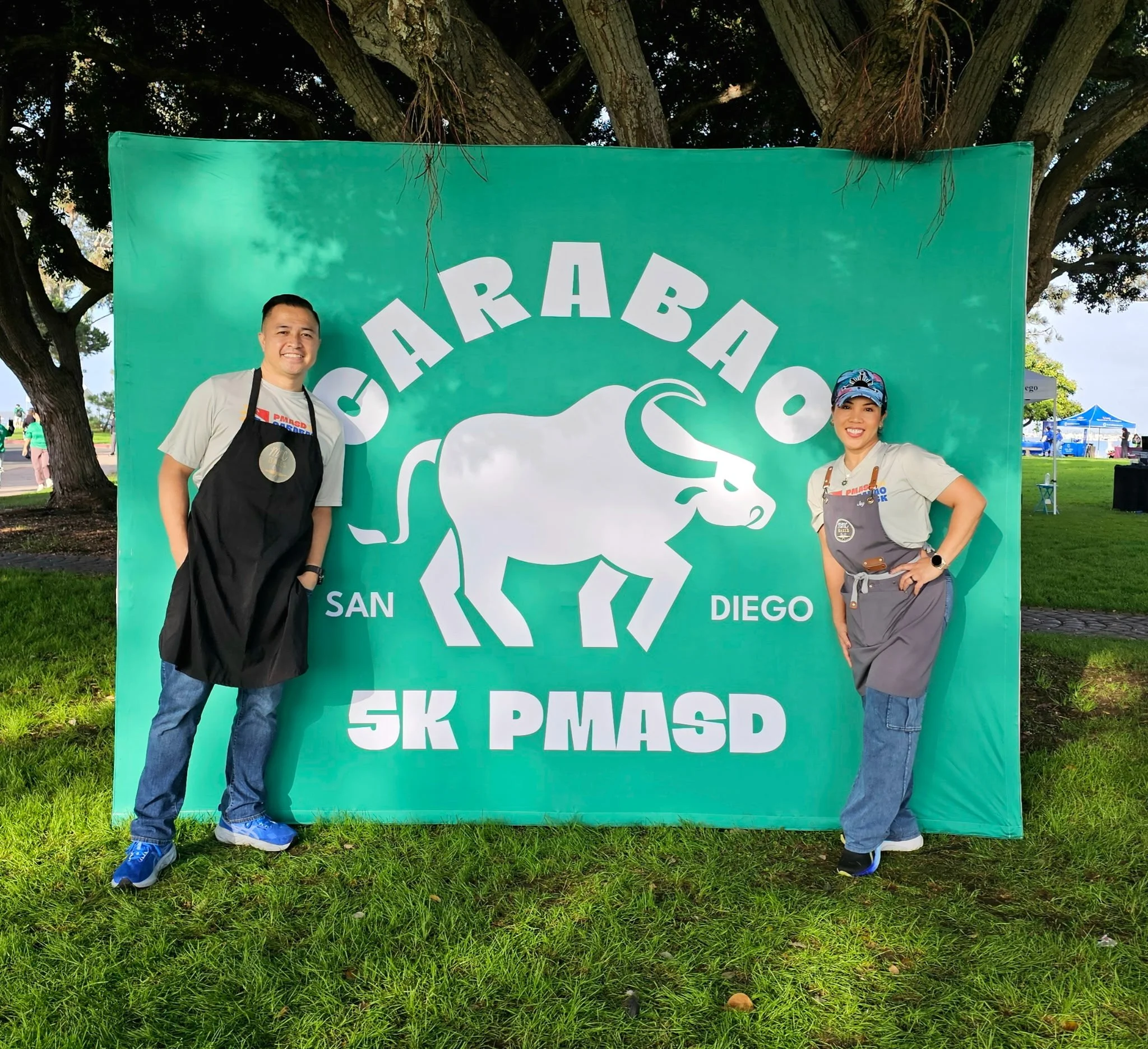 Two people standing outdoors representing the MMC Bakes Chula Vista business in front of a green backdrop with a rhinoceros graphic and text. PMA Carabao 5K event. They are smiling and posing for the photo.