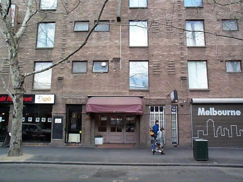 A brick apartment building with corner entrance and a maroon awning, pedestrians walking on sidewalk, leafless tree, and a mural with the word 'Melbourne' on a closed storefront.