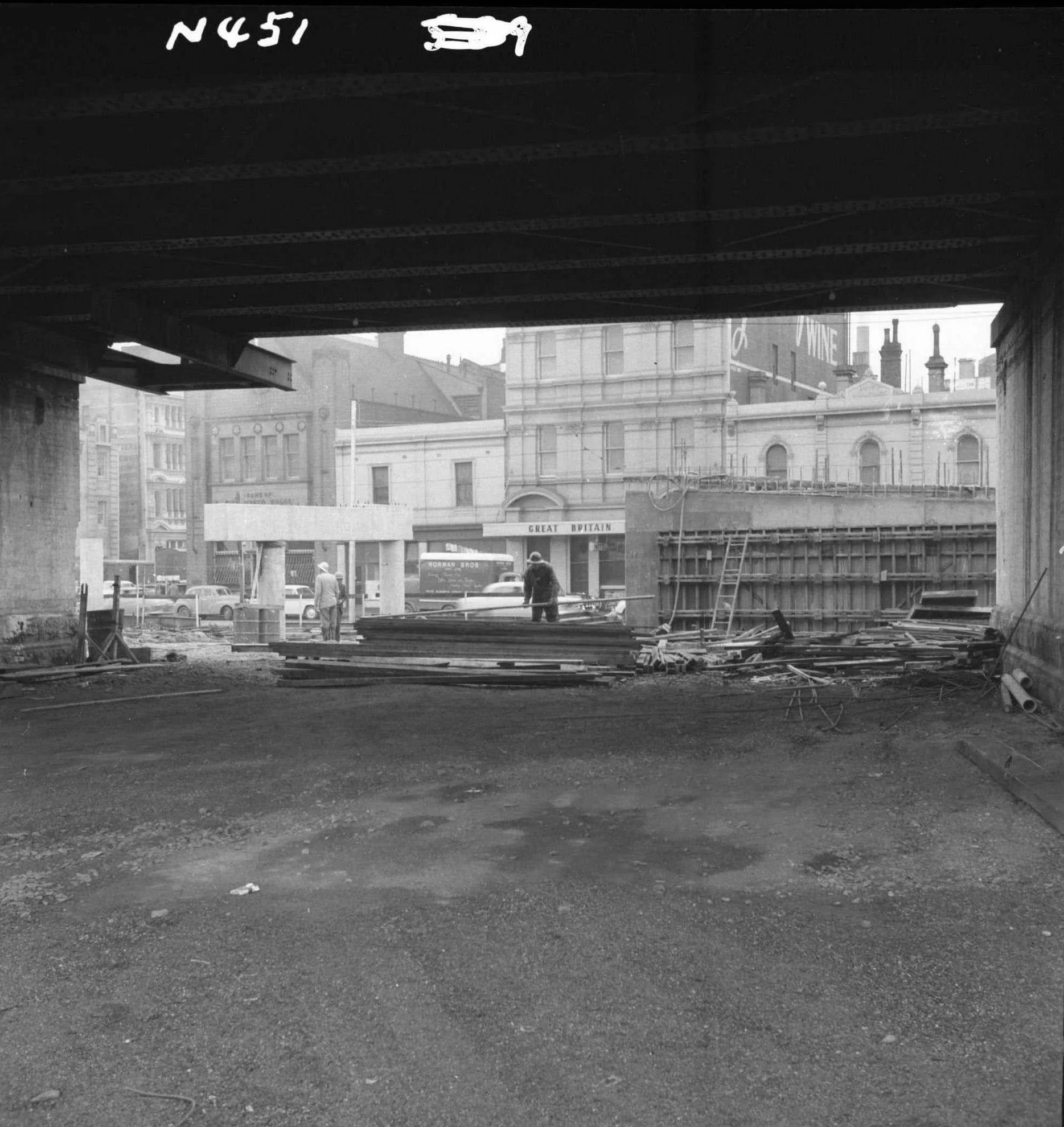 Black and white photo of construction site with a large opening in a building, showing multiple workers and a bus in the background, and buildings with billboards and chimneys in the distance.