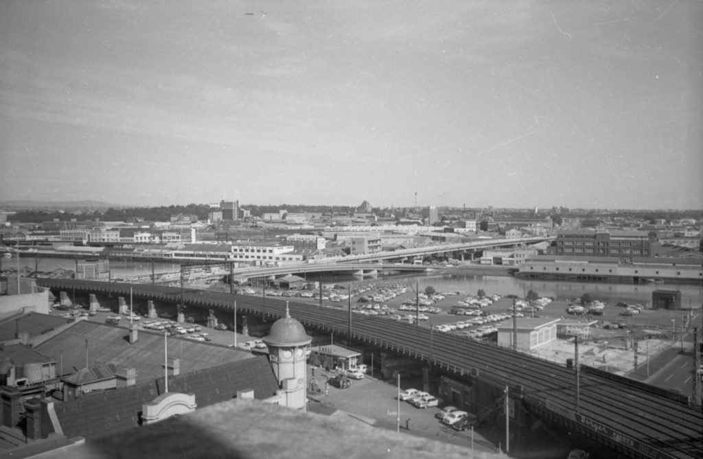 Black-and-white photo of a cityscape with train tracks and a parking lot in the foreground, multiple buildings, and a waterway in the distance.