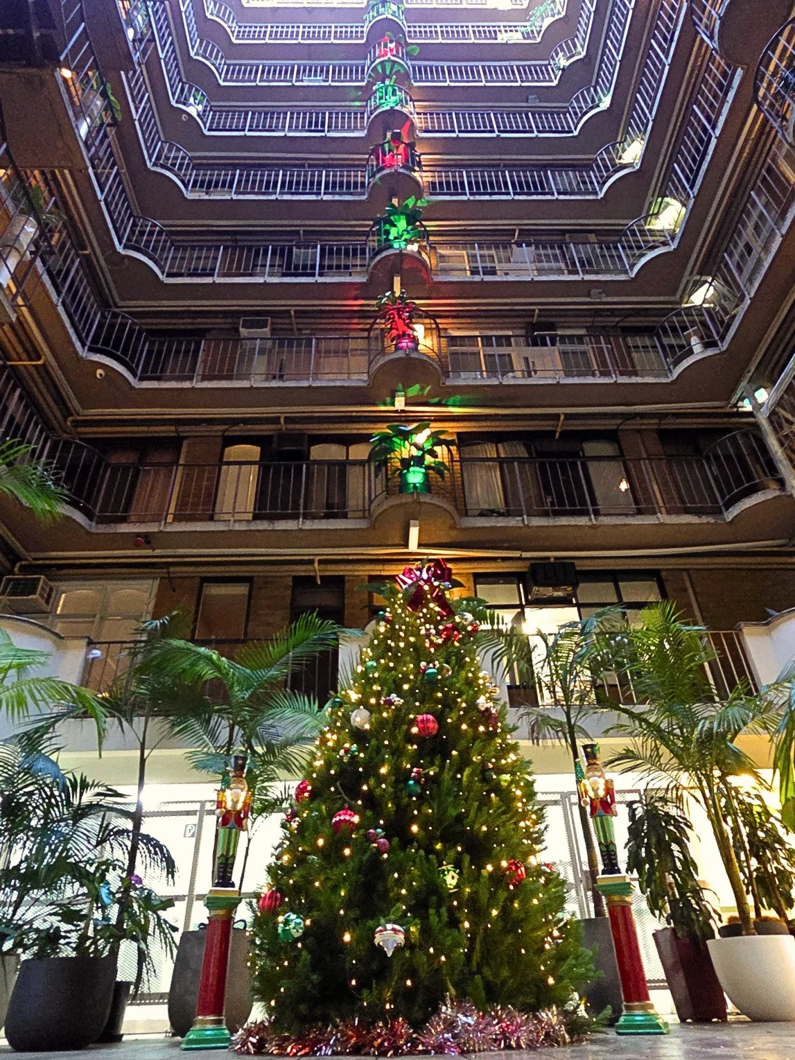 Indoor hotel atrium decorated for Christmas with a Christmas tree in center, lights, ornaments, and decorations, surrounded by potted plants, with multiple floors visible and holiday light decorations hanging from upper balconies.