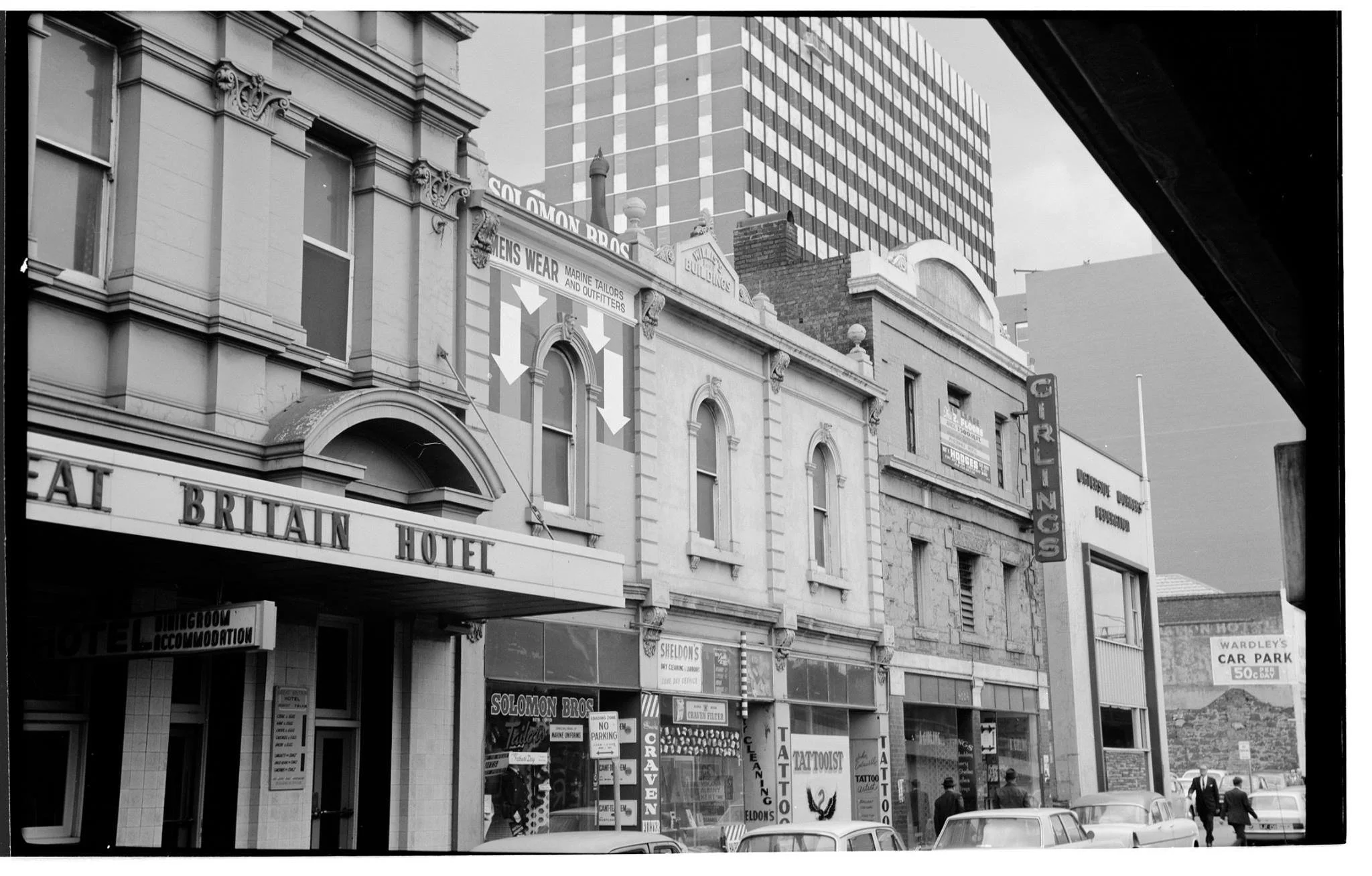 Black and white photo of a city street with historic buildings and modern high-rise in the background. The street level has various storefronts, including a hotel called the 'Britain Hotel' and signs for tattoo parlors, car parking, and other busines
