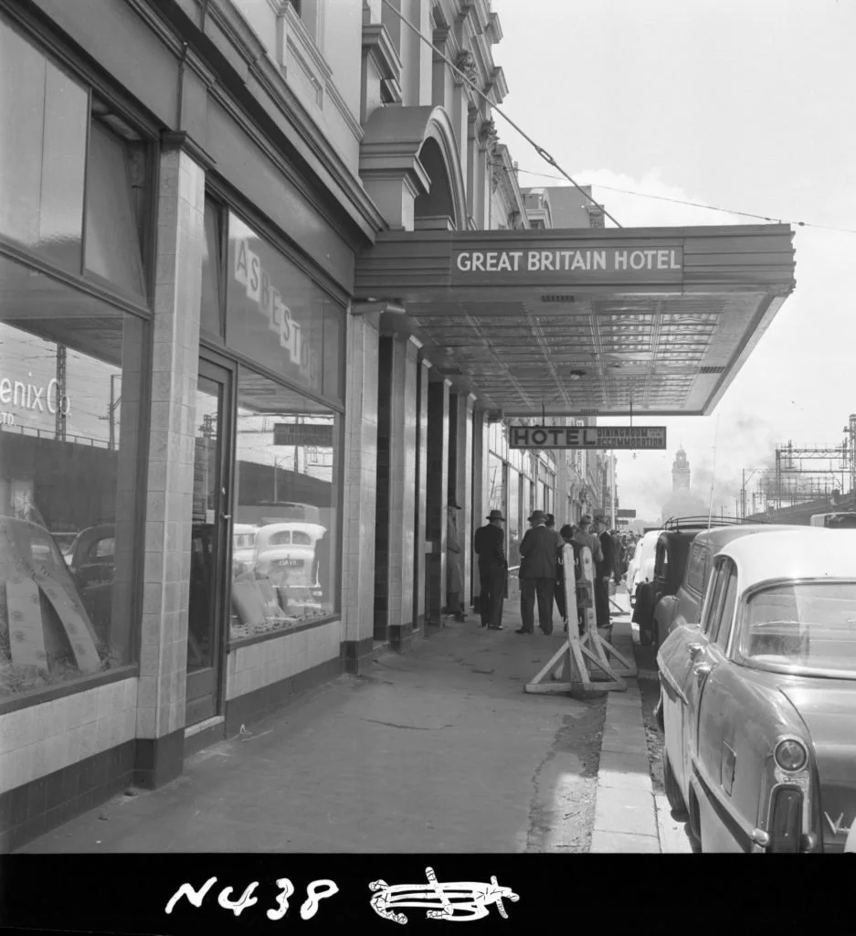 Black and white photo of the sidewalk outside the Great Britain Hotel, with a marquee sign and a group of men standing near vintage cars parked along the street. The architecture features large windows and ornate building styles.