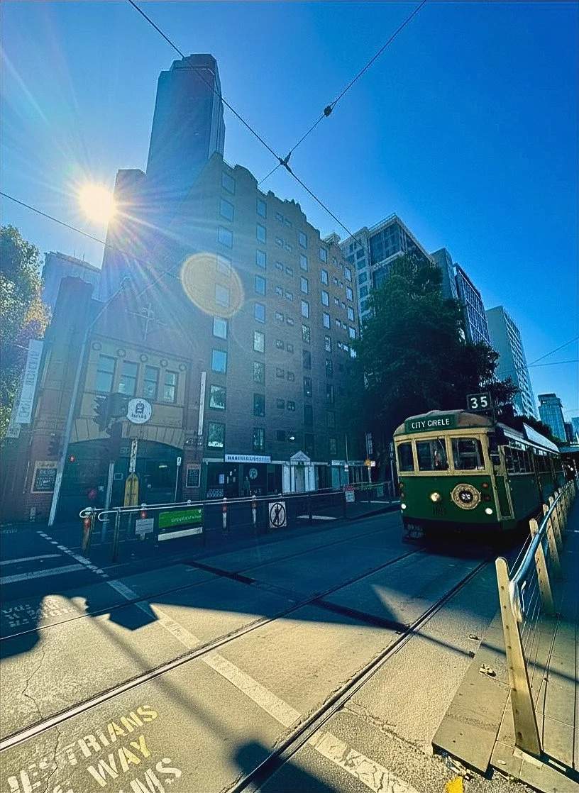 A city street with a green and cream streetcar labeled "CITY CIRCLE," overhead tram wires, a tall modern building, and a clear blue sky with the sun shining brightly, creating lens flare.