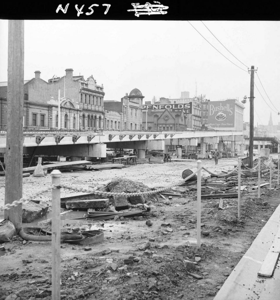 A black and white photo of a street under construction with old buildings in the background. Construction materials and a fallen turtle are on the dirt ground, with a rope fence in the foreground. Power lines and signs, including "OPENFOLDS" and "Bus