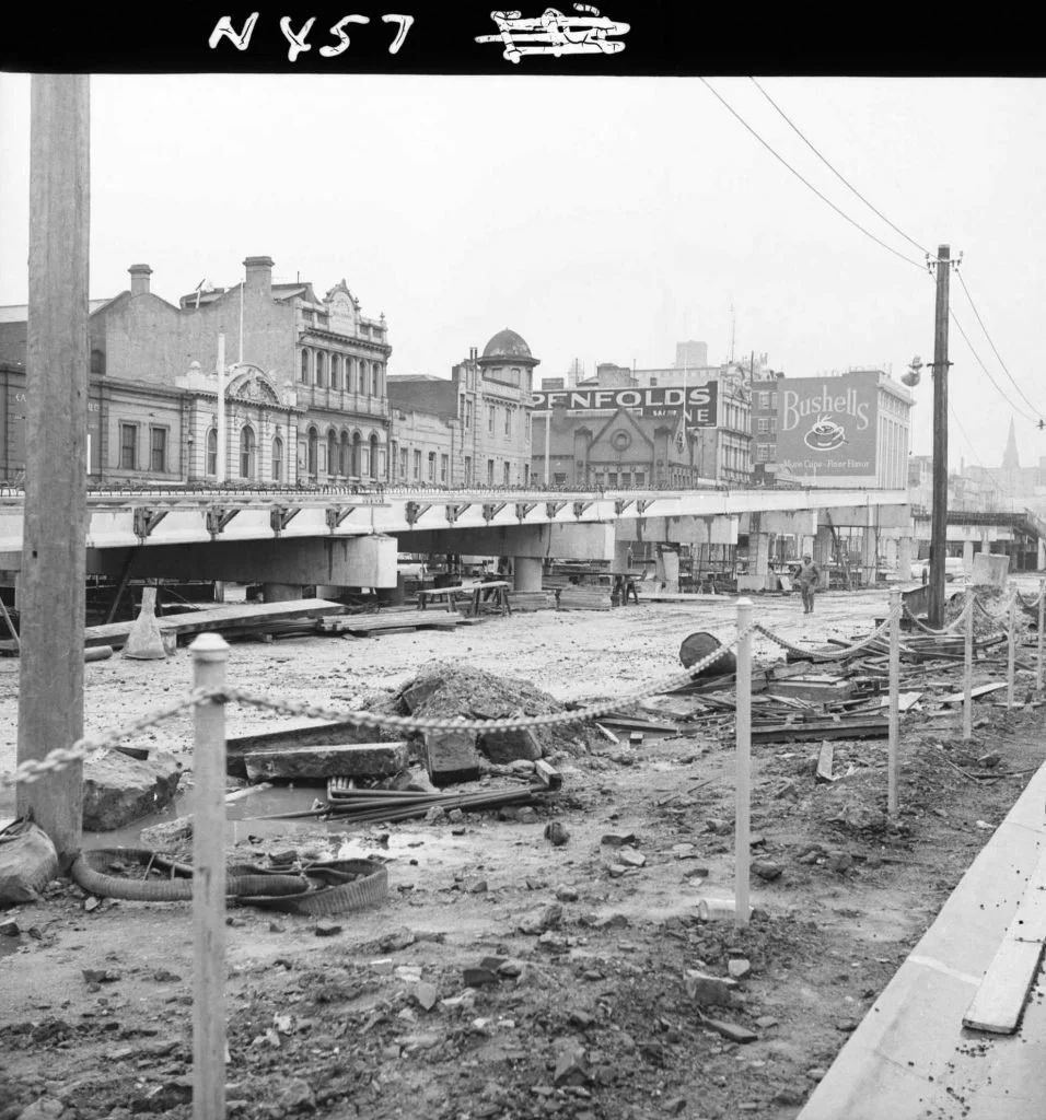 Black and white photo of an under-construction bridge on a city street, with old buildings and billboards in the background; construction debris and a worker are visible in the foreground.