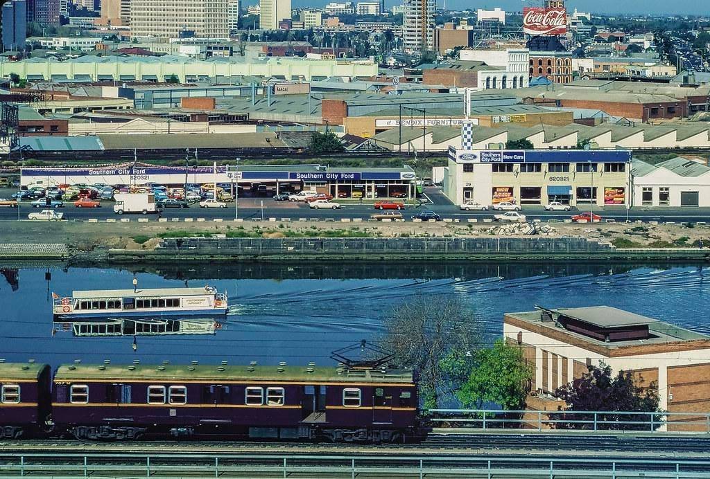 Cityscape with a river, a boat, train, and buildings including a Coca-Cola billboard.
