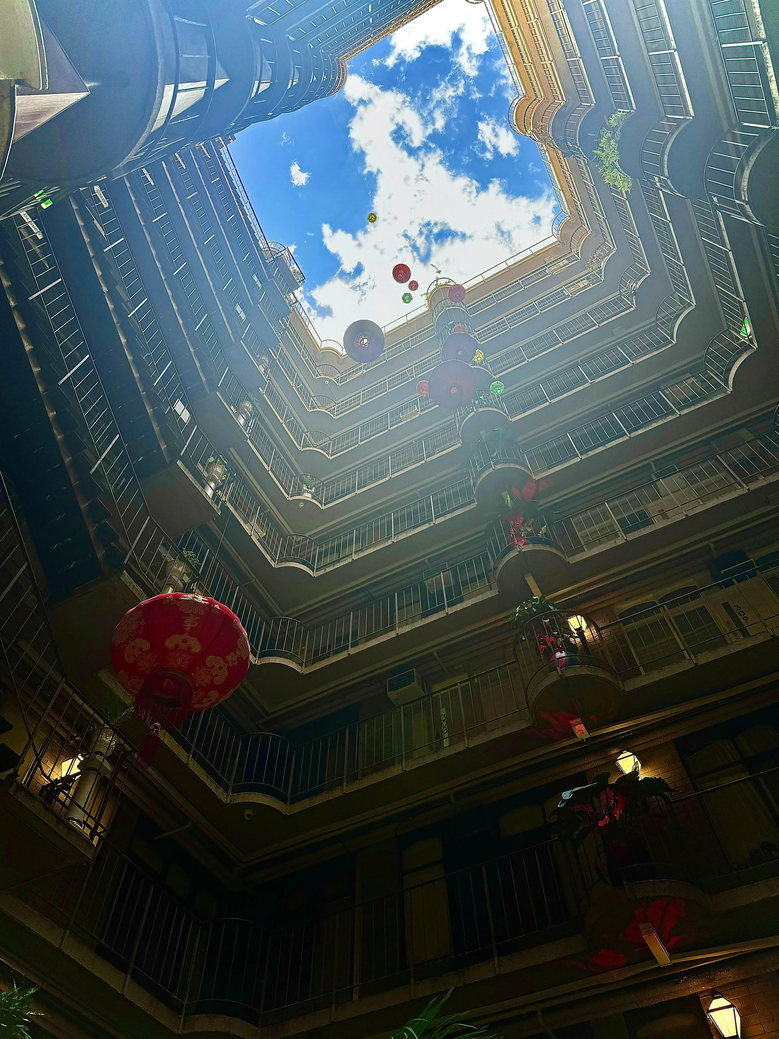 A view looking up from the courtyard of a multi-story building with balconies on each level, balloons and lanterns hanging, and a partially cloudy blue sky visible at the top.