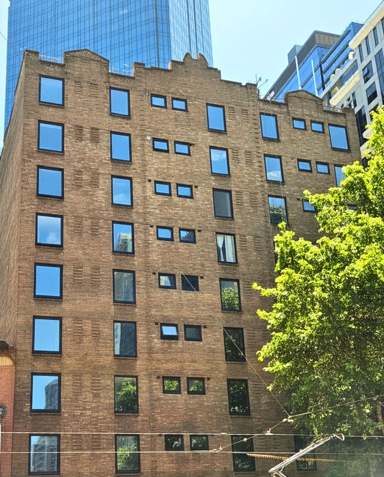 A multi-story brick building with various-sized windows, surrounded by taller modern glass and concrete buildings, and a leafy green tree in the foreground.