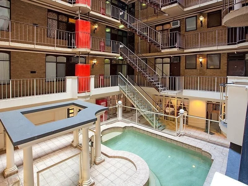 Indoor hotel courtyard with a small pool, multiple floors with balconies, red fire escape stairs, brick walls, and glass-paneled barriers.