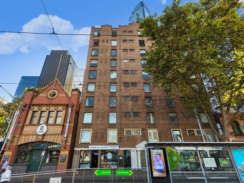 Tall brown brick apartment building with multiple windows, a tree on the right, and nearby storefronts at street level in an urban area.