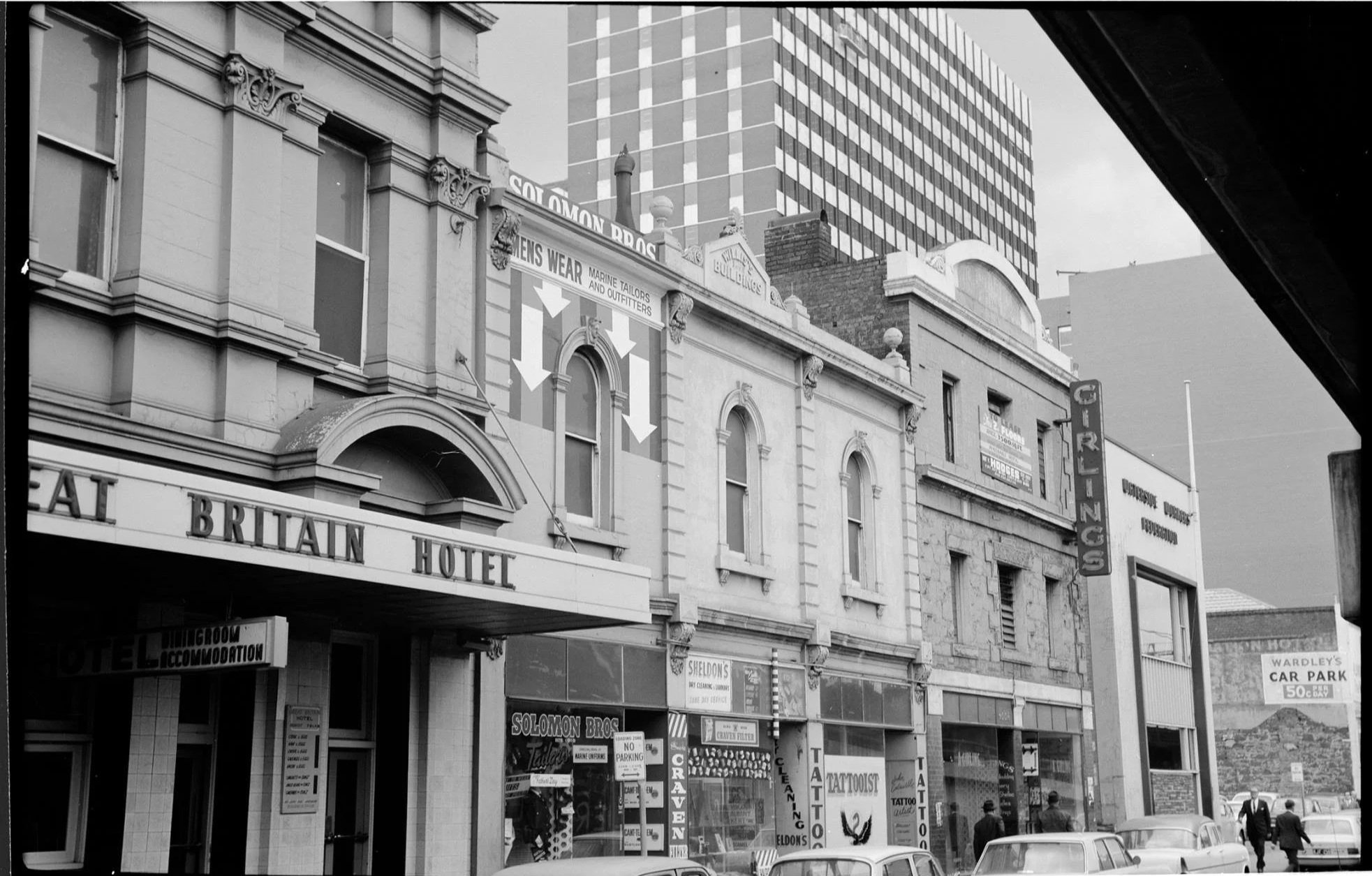 Black and white photo of a city street with a theater marquee for the British Hotel and various storefronts, signs for Solomon Bros, tattoo parlor, and other businesses, pedestrians walking on the sidewalk, parked cars in front, and tall buildings in