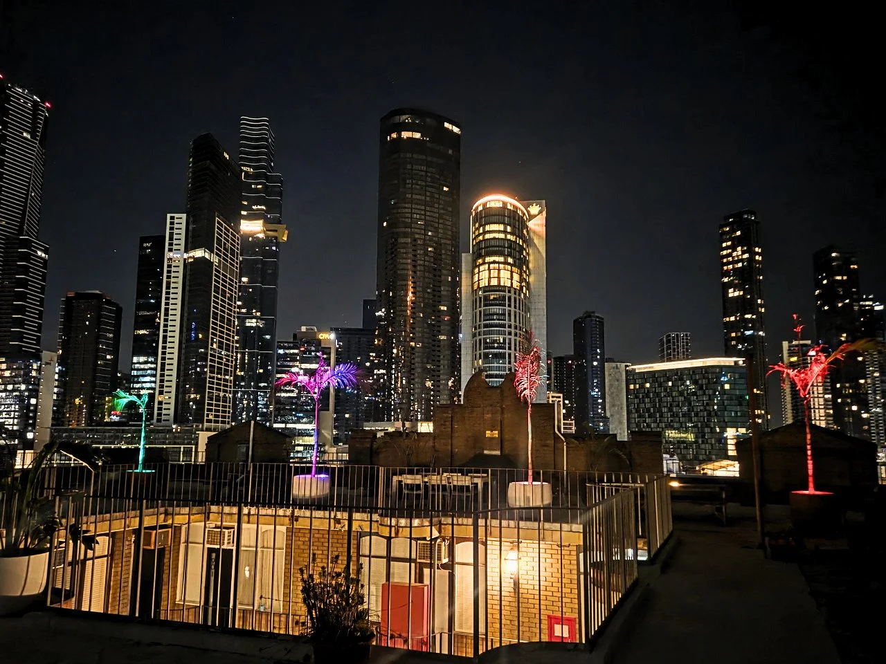 Night view of a city skyline with illuminated skyscrapers and colorful neon palm tree lights on a rooftop or terrace.