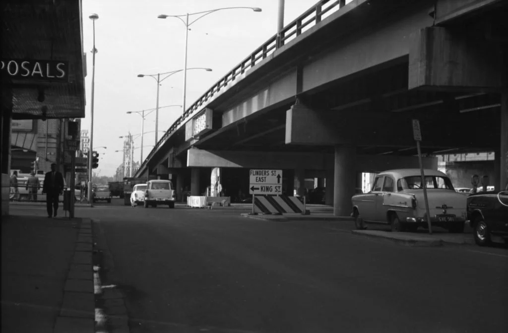 Black and white photo of a city street scene. An elevated highway runs above the street, with cars parked underneath. A pedestrian is walking on the sidewalk near a bus stop, and a street sign points to Flinders Street East and King Street.