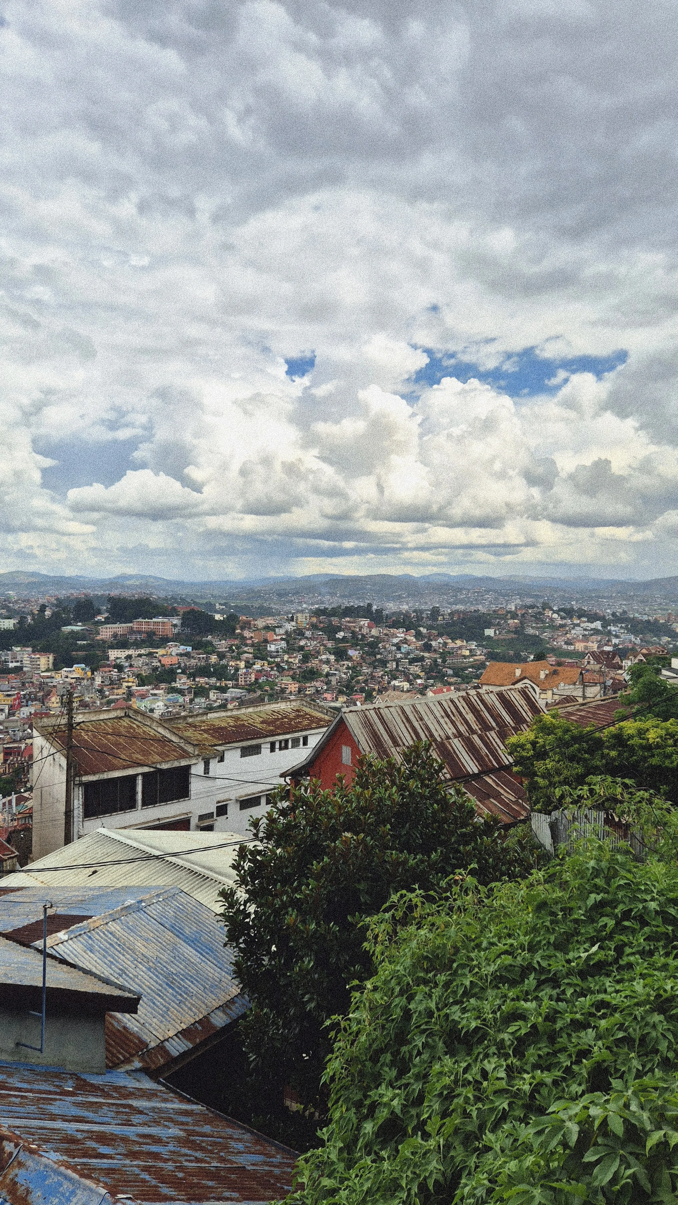 Vue panoramique d'un quartier résidentiel sur une colline avec des toits en tôle rouillée, des maisons colorées et des arbres verts sous un ciel nuageux.
