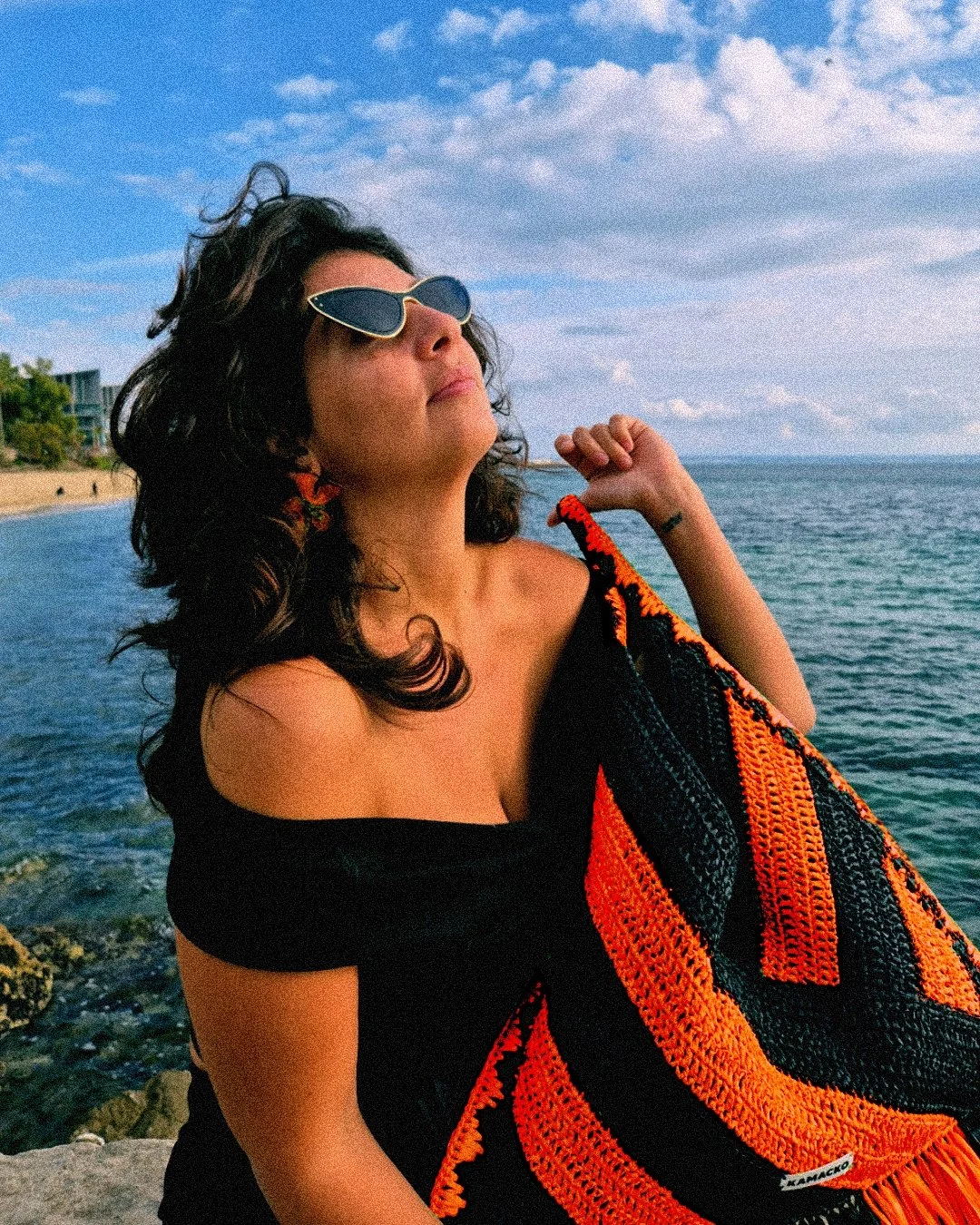 Woman by the sea holding the crochet bag Le Agulla from the brand KAMACKO. The bag is bold, graphic, with black and orange large stripes handmade in raffia.