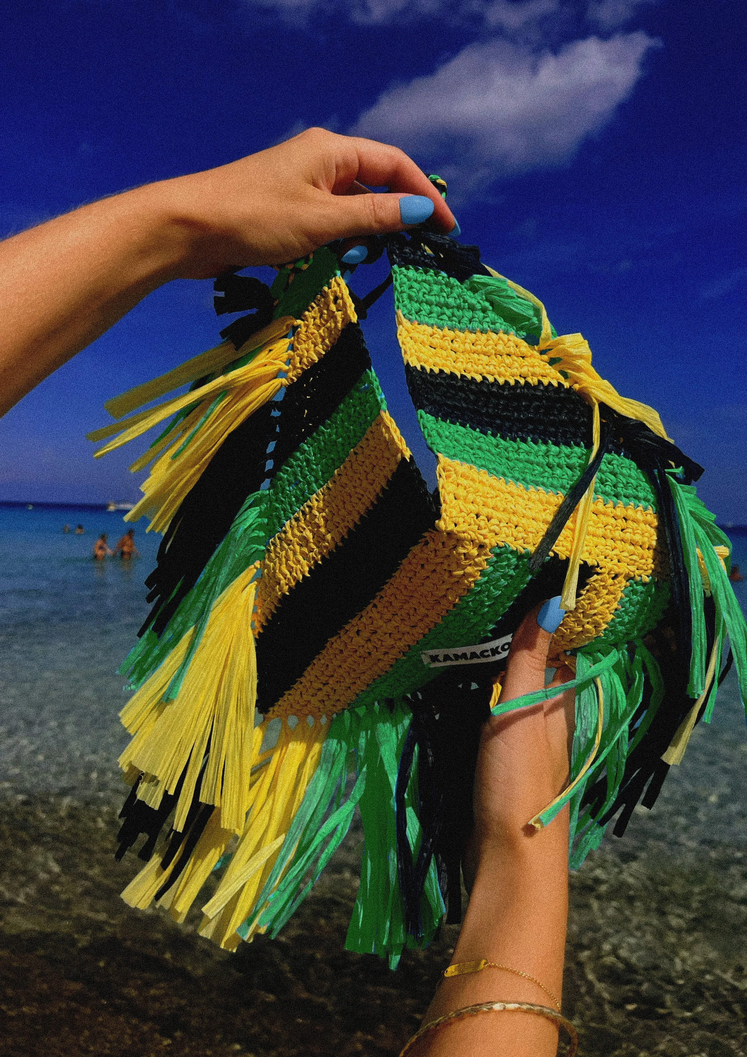 Hands are holding the bag Le Deià from KAMACKO. Graphic colourful bold handmade crochet bag in raffia with black yellow and green stripes with raffia fringes. In the background you see blue sky and the sea of Cala Agulla in Mallorca.