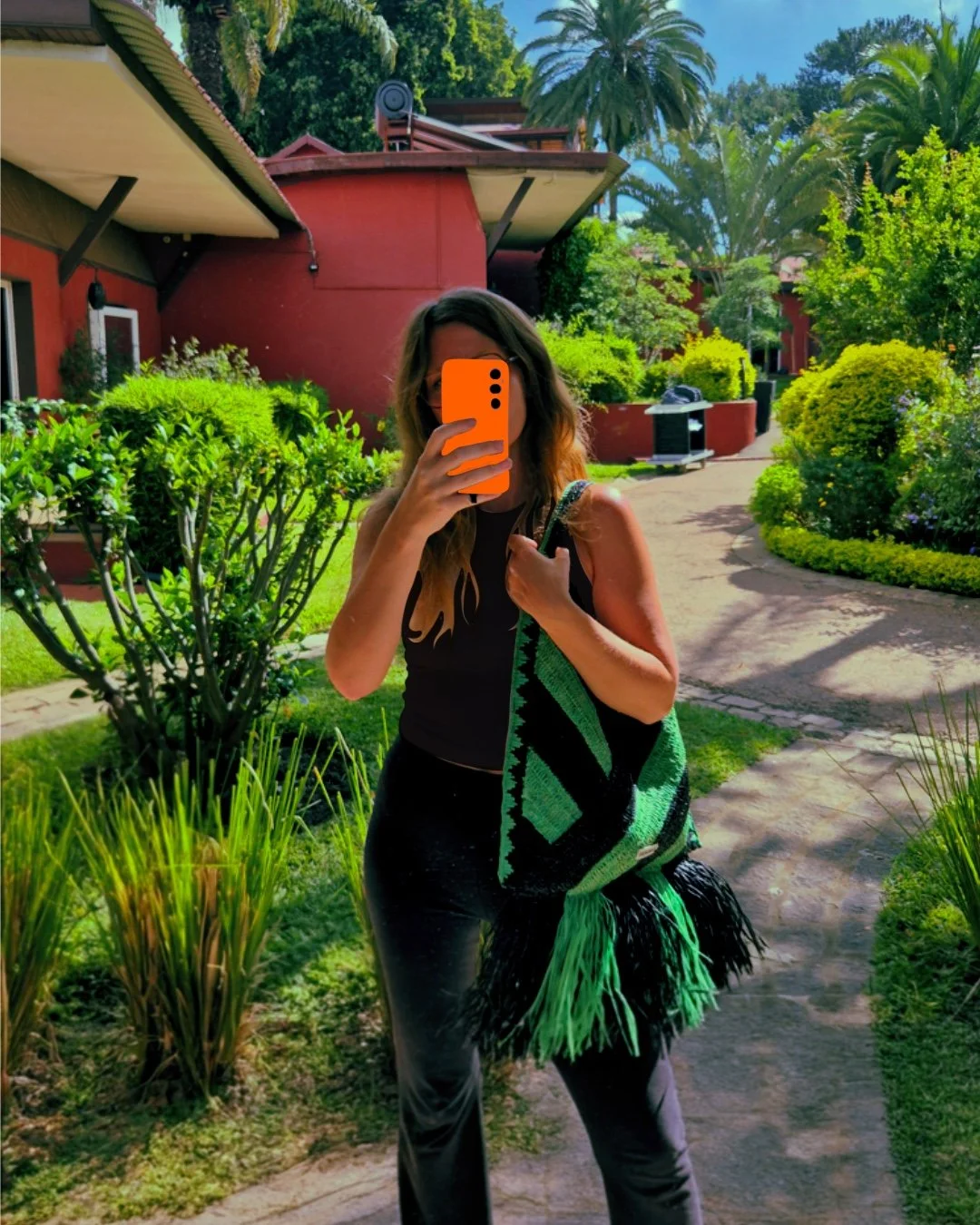Woman doing a selfie in a mirror holding the crochet bag Le Agulla from the brand KAMACKO. The bag, handmade crochet in raffia, has large bold and graphic black and green stripes with raffia fringes. The back ground has green vegetation.