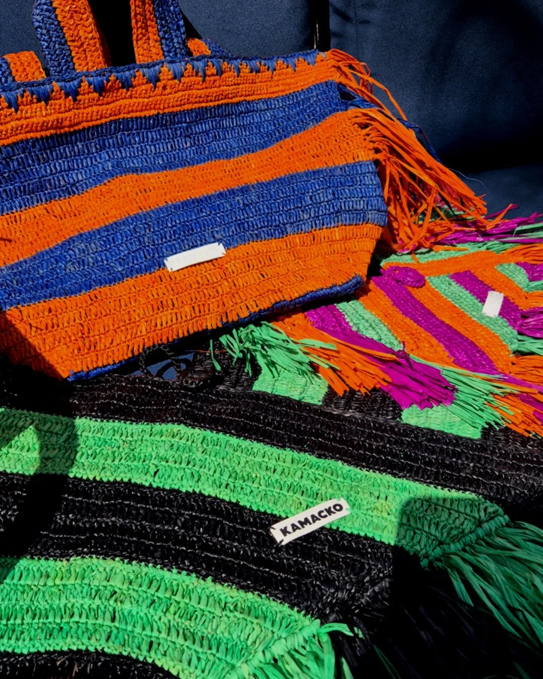 Colourful graphic bold stripes crochet bags in raffia from the brand Kamacko. You can see Le Deià in stripes green orange and purple, Le Sóller in stripes blue and orange and Le Agulla in stripes black and green. You can see the logo tag "KAMACKO".