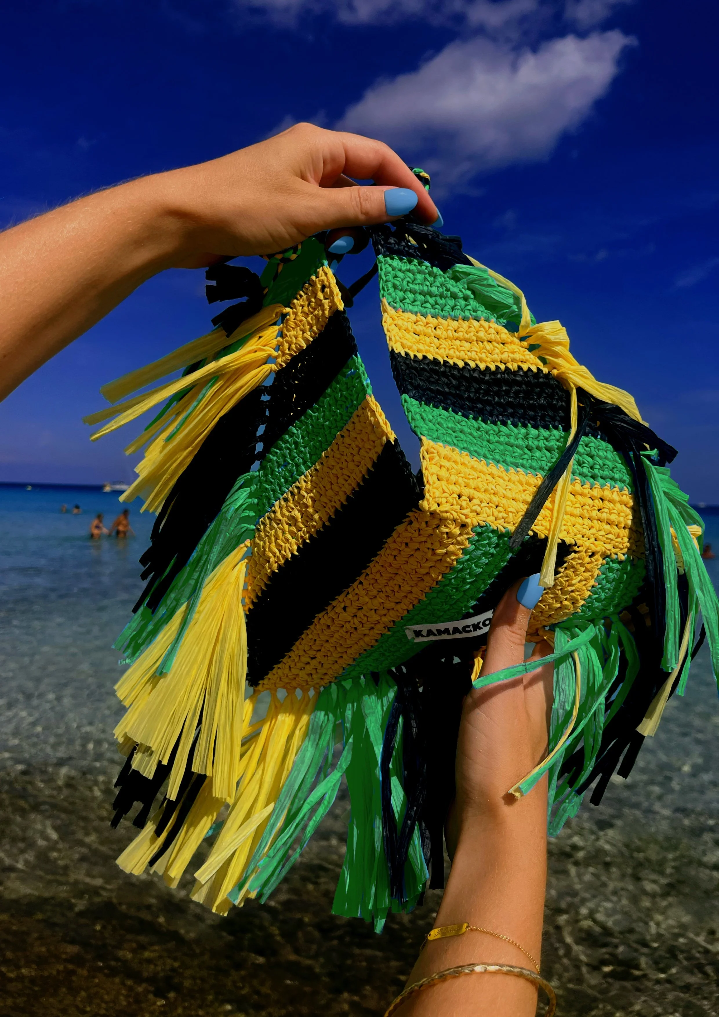 Hands are holding the bag Le Deià from KAMACKO. Graphic colourful bold handmade crochet bag in raffia with black yellow and green stripes with raffia fringes. In the background you see blue sky and the sea of Cala Agulla in Mallorca.